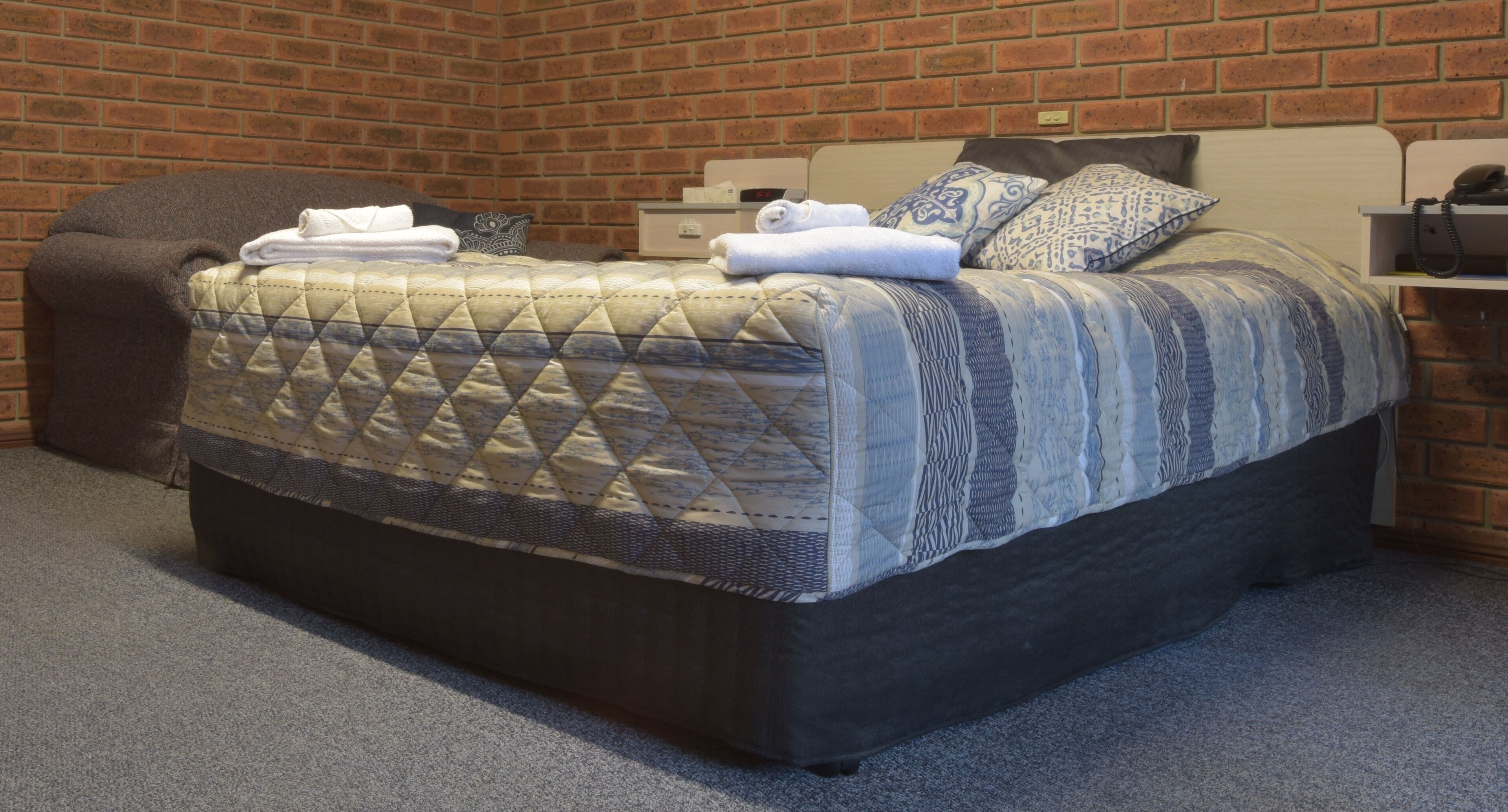A neatly made bed with a quilted cover, a bedside table with a lamp, and a brick wall background.