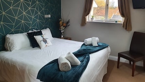 A neatly made bed with white linens, teal throw pillows, and a teal blanket. There is a chair to the right of the bed and a window with curtains. - The Nags Head (Norwich)