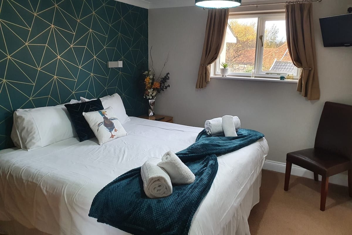 Un lit bien fait, avec du linge de lit blanc, des coussins turquoise et une couverture turquoise. Il y a une chaise à droite du lit et une fenêtre avec des rideaux.