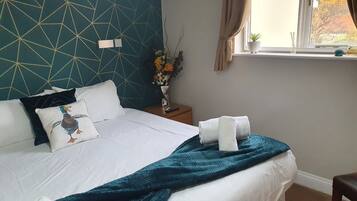 Una cama bien hecha con un edredón y almohadas color turquesa, un florero con flores y una ventana con cortinas.