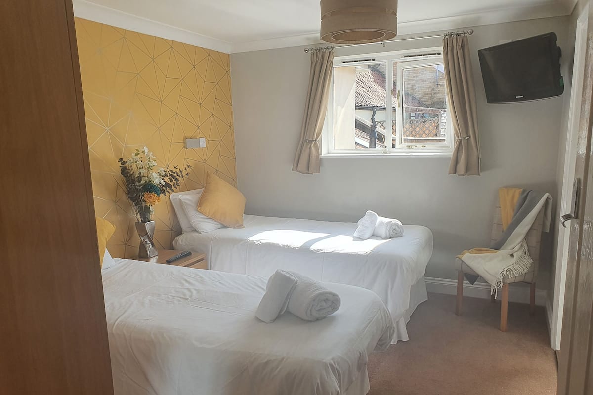 Une chambre d’hôtel avec deux lits, un téléviseur fixé au mur, une fenêtre avec des rideaux et une chaise avec une couverture.