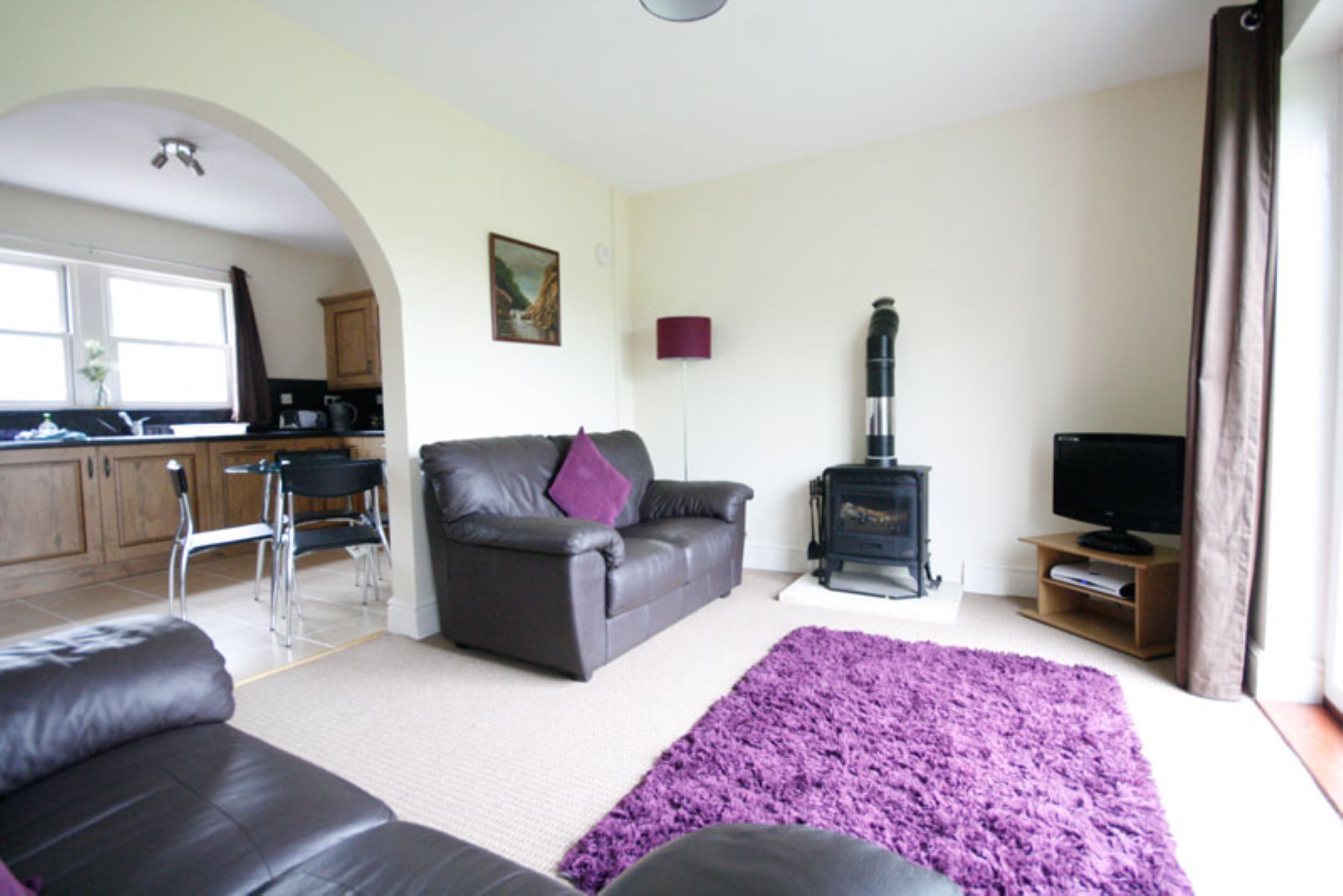 A living room with a sofa, a coffee table, a fireplace, a television, and a kitchen area with cabinets and a window.
