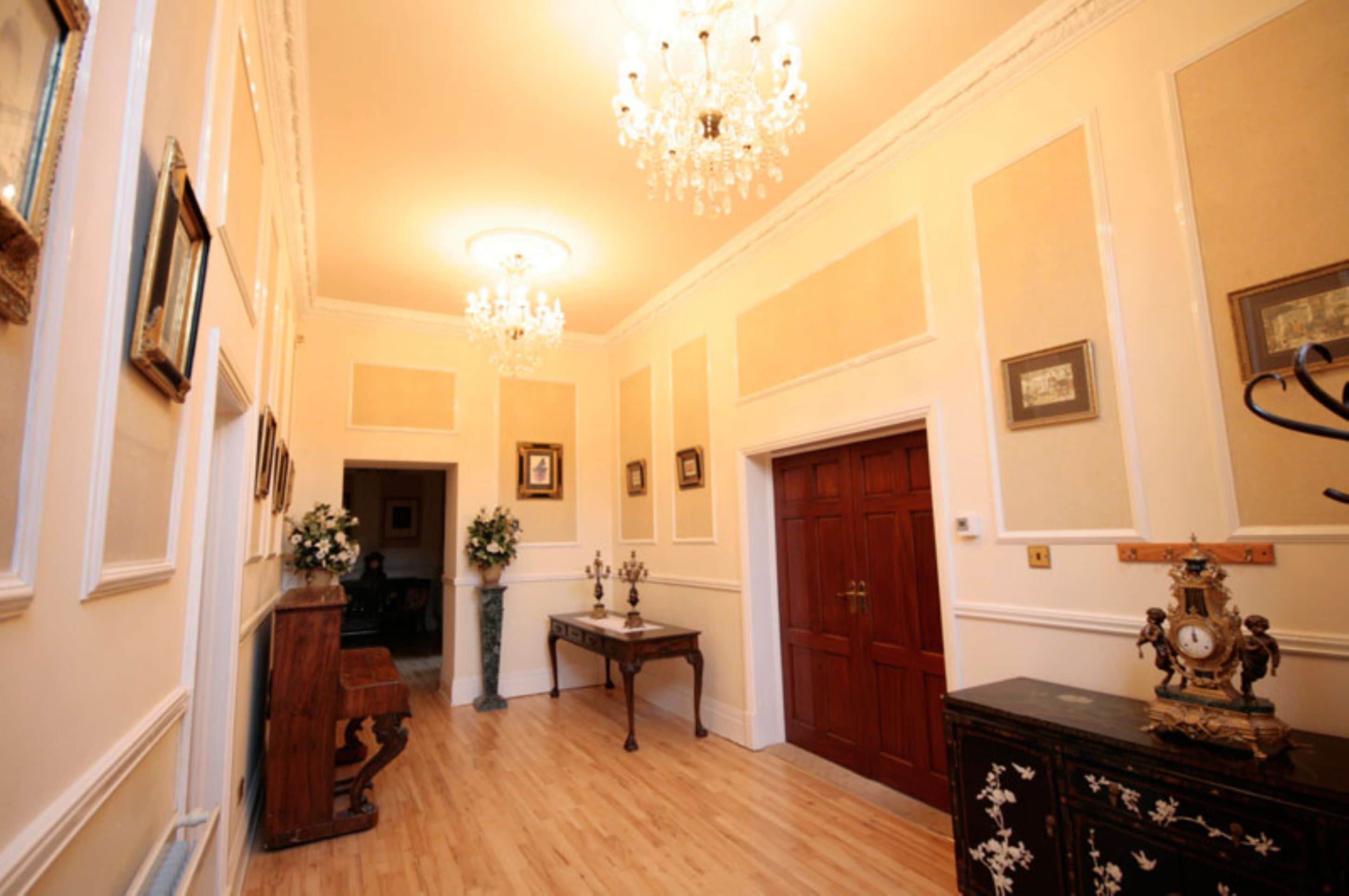 A hallway with wooden flooring, a chandelier, framed pictures on the walls, and a wooden door.