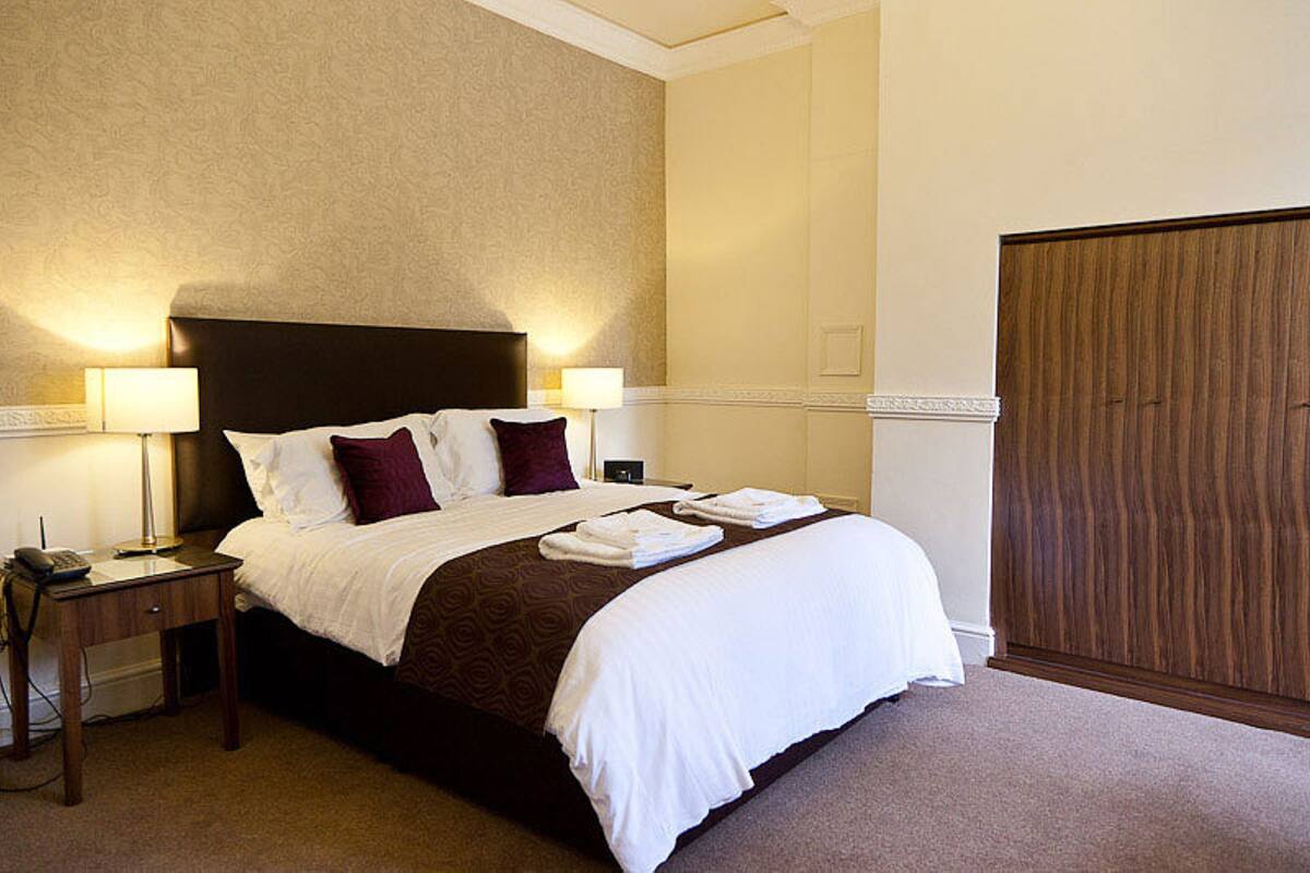 A hotel room with a large bed, two bedside lamps, a wooden wardrobe, and a carpeted floor.