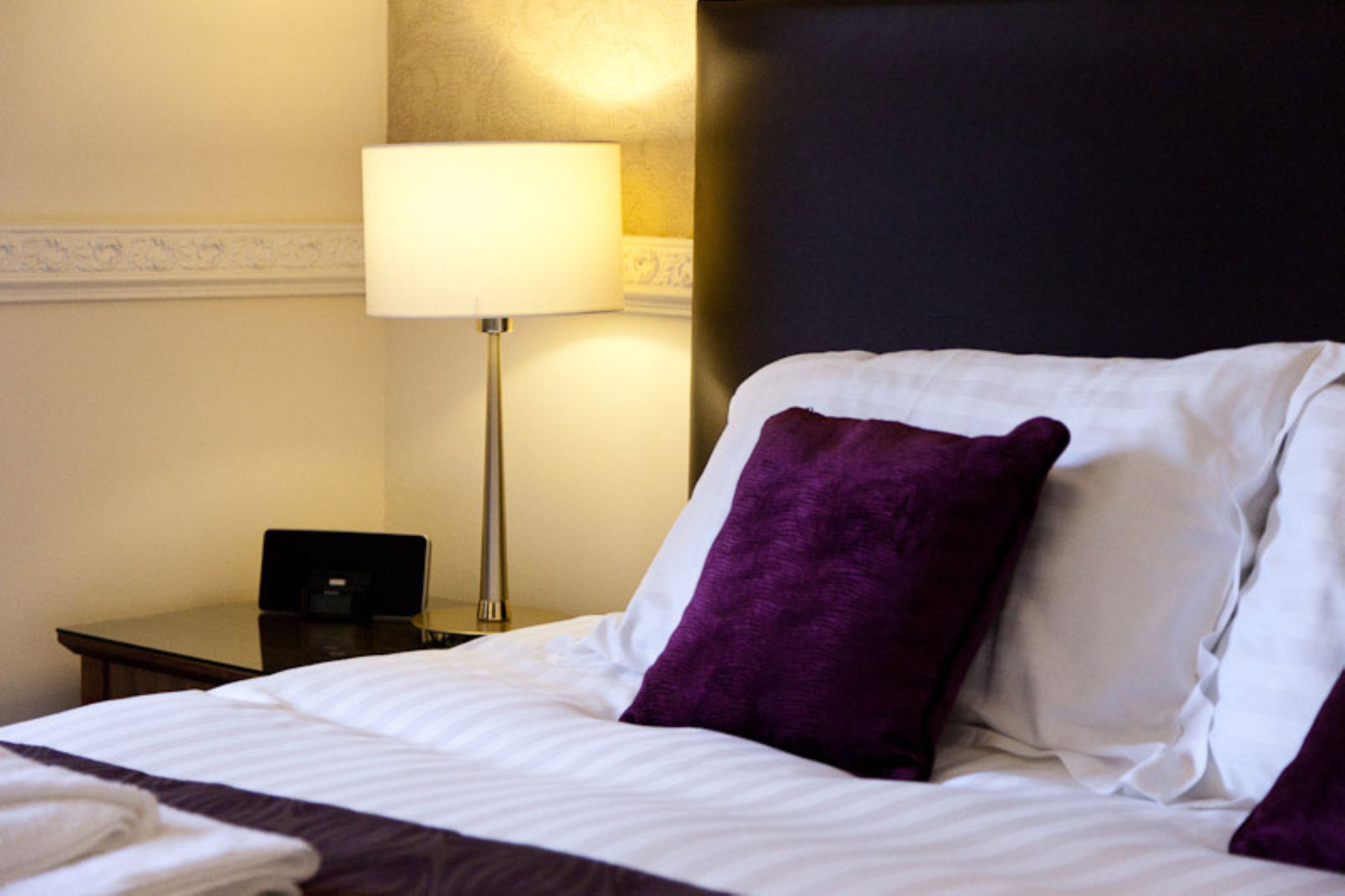 A hotel room with a neatly made bed, a bedside table with a lamp, and a dark headboard.