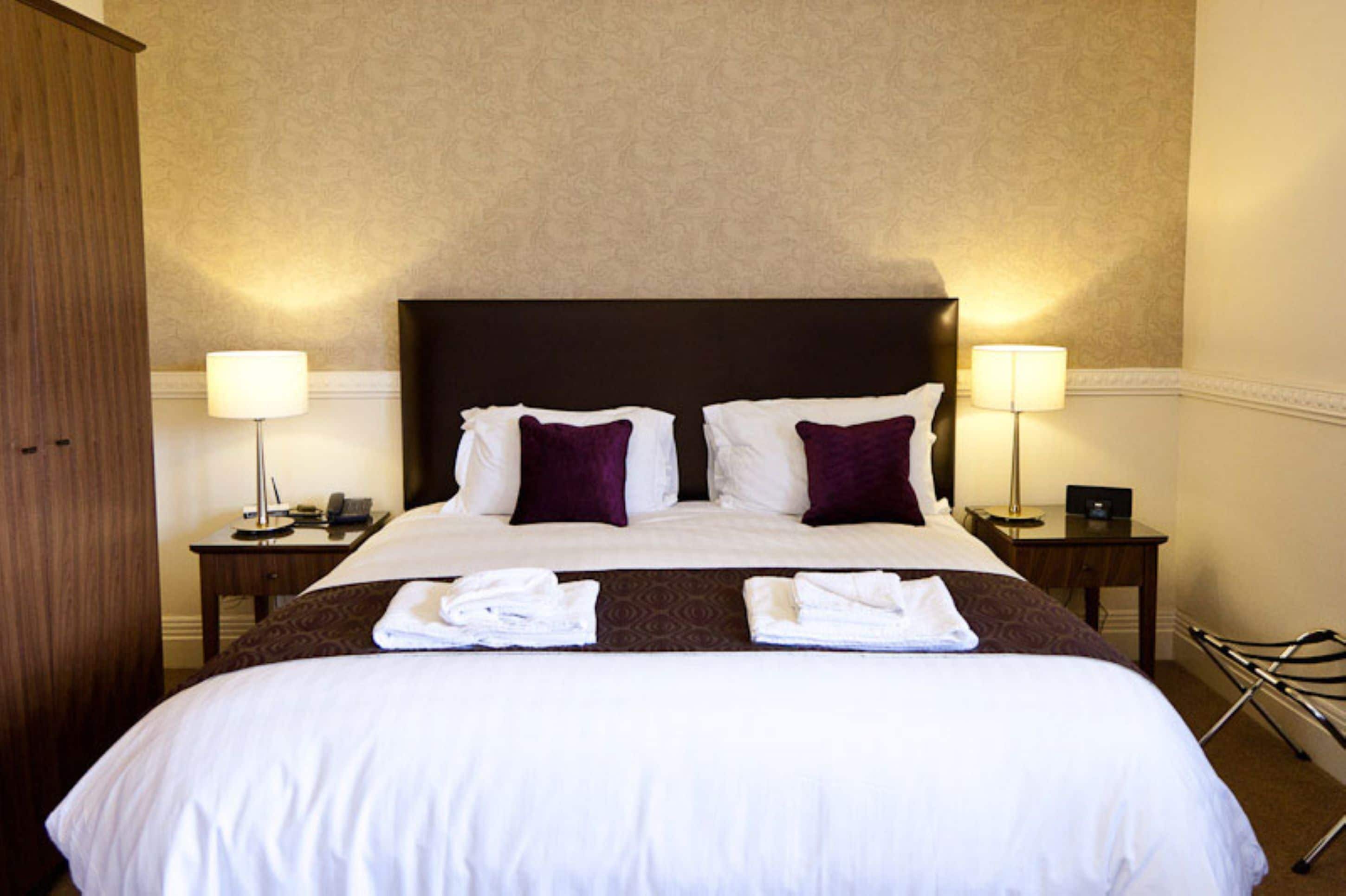Cama bem arrumada com travesseiros brancos e roxos, duas mesas de cabeceira com abajures e uma toalha dobrada na cama.