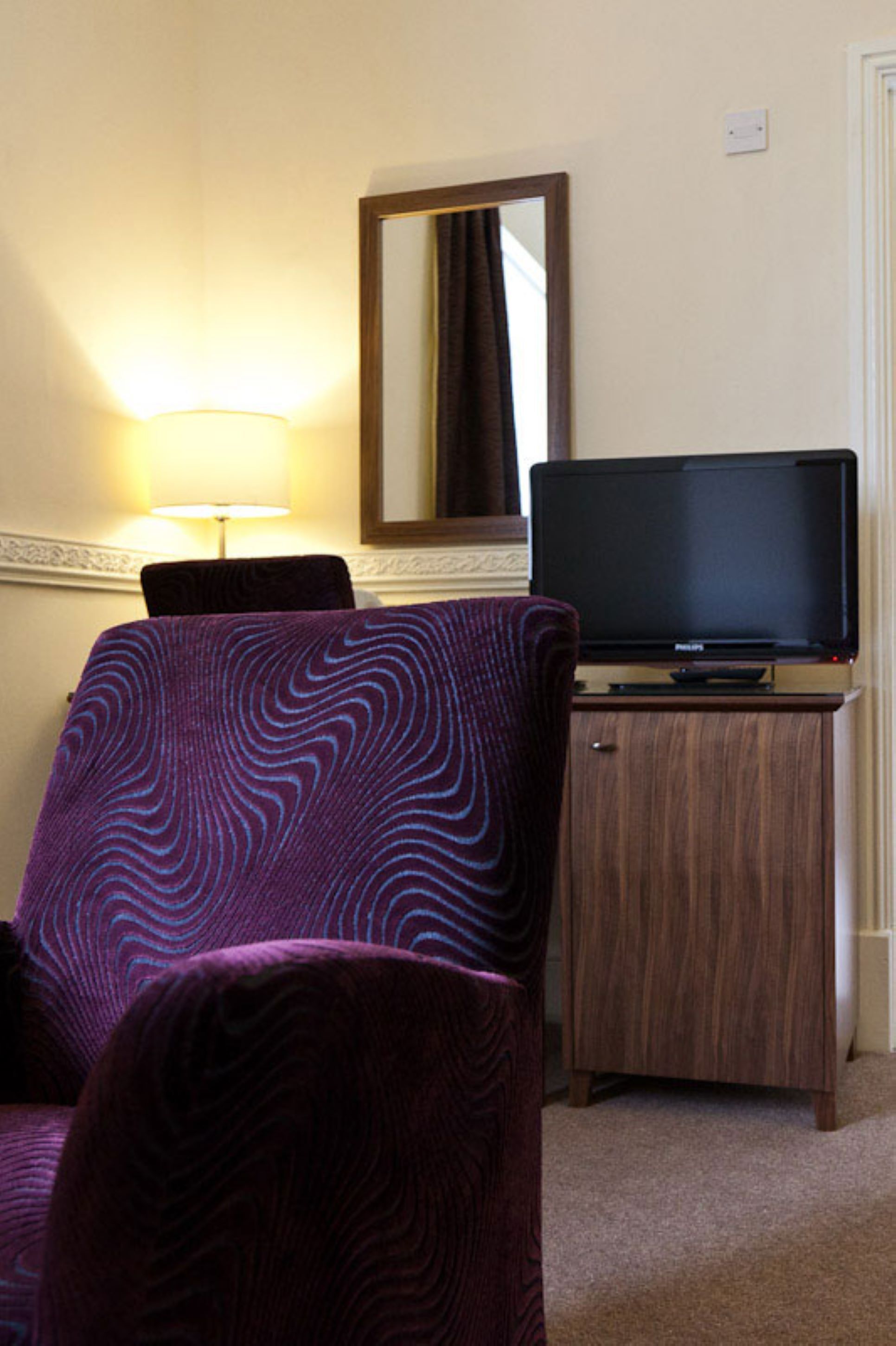 Une chambre d’hôtel avec un fauteuil à motifs violets, un meuble en bois sur lequel est posée une télévision, et un miroir fixé au mur.