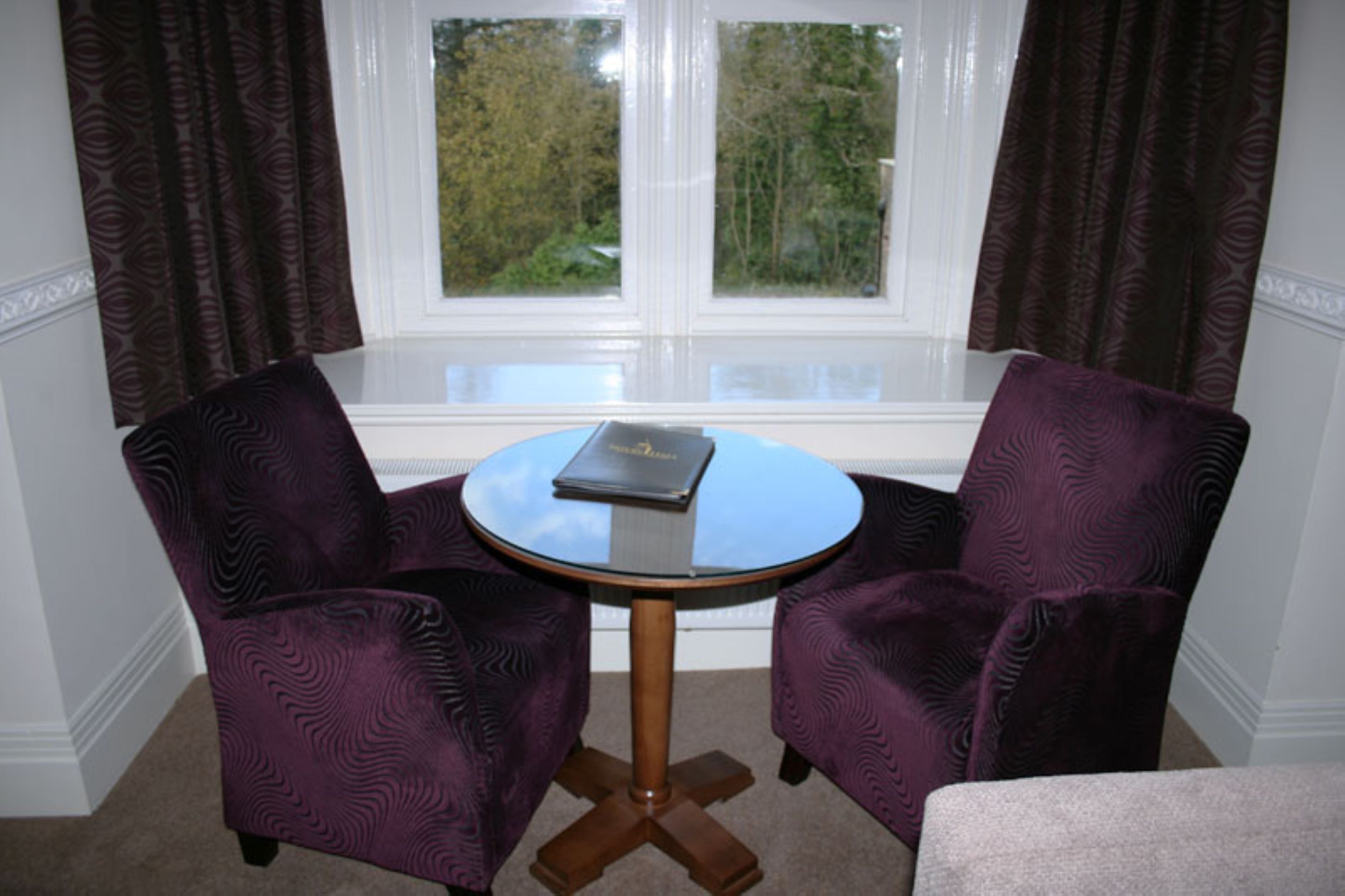 A room with two purple upholstered chairs around a small round table with a book on it, near a window with curtains.