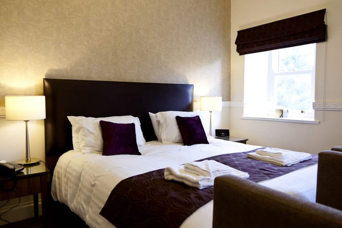 A neatly made bed with white and purple bedding, a dark headboard, and a window with a view of trees.