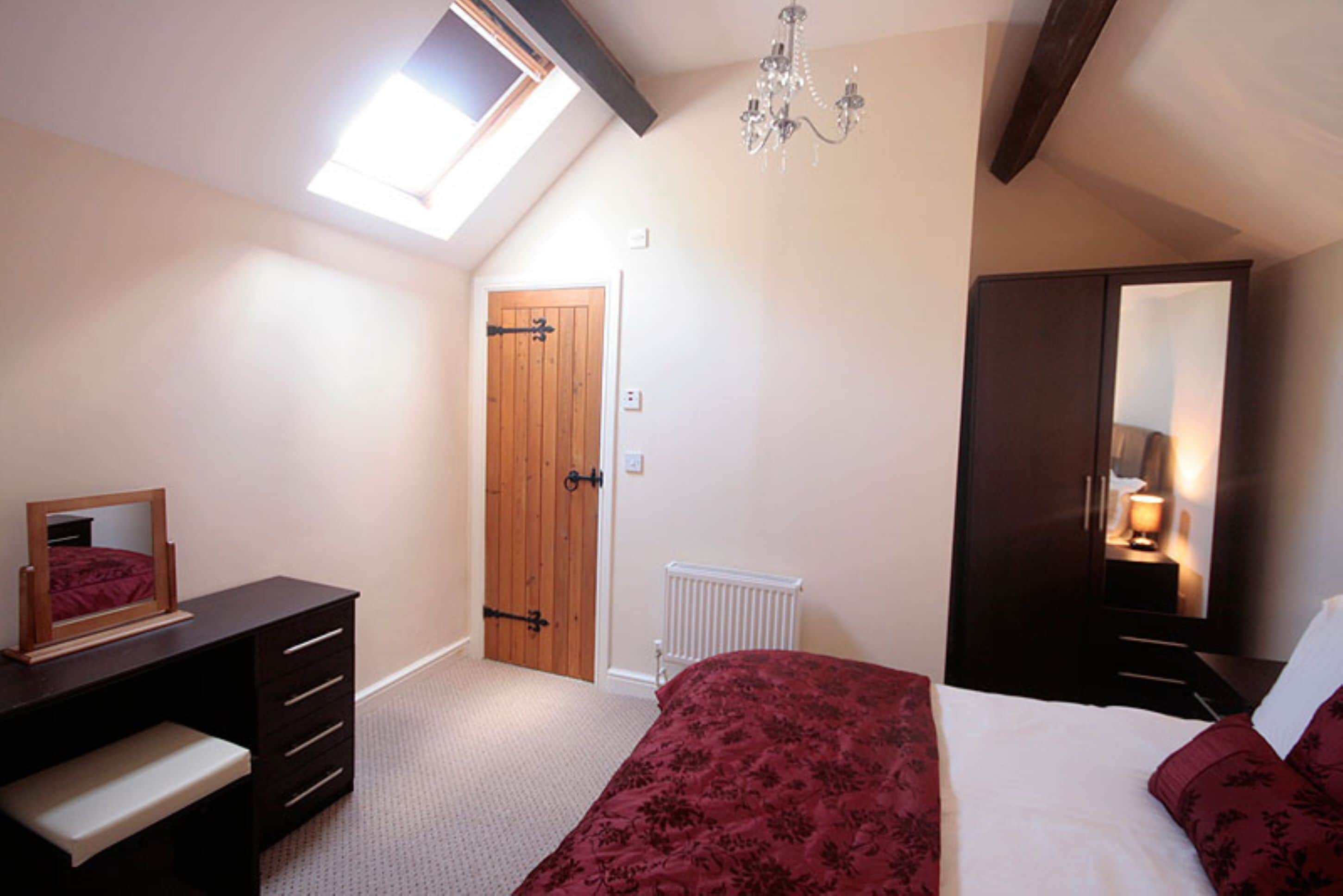 A bedroom with a sloped ceiling, a skylight, a wooden door, a radiator, a bed with a red bedspread, a dresser, and a mirror.