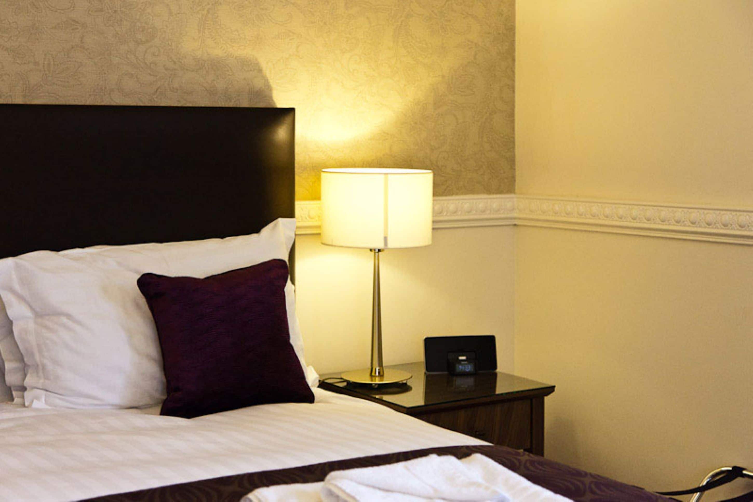 A neatly made bed with white and dark purple pillows, a bedside lamp, and a small nightstand with a clock.