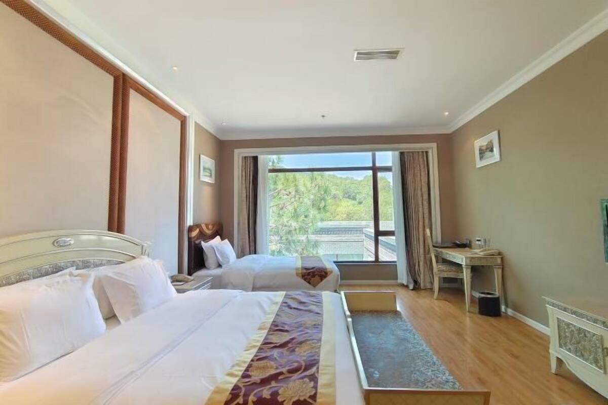 A hotel room with two beds, a desk, a chair, and a large window with a view of greenery.