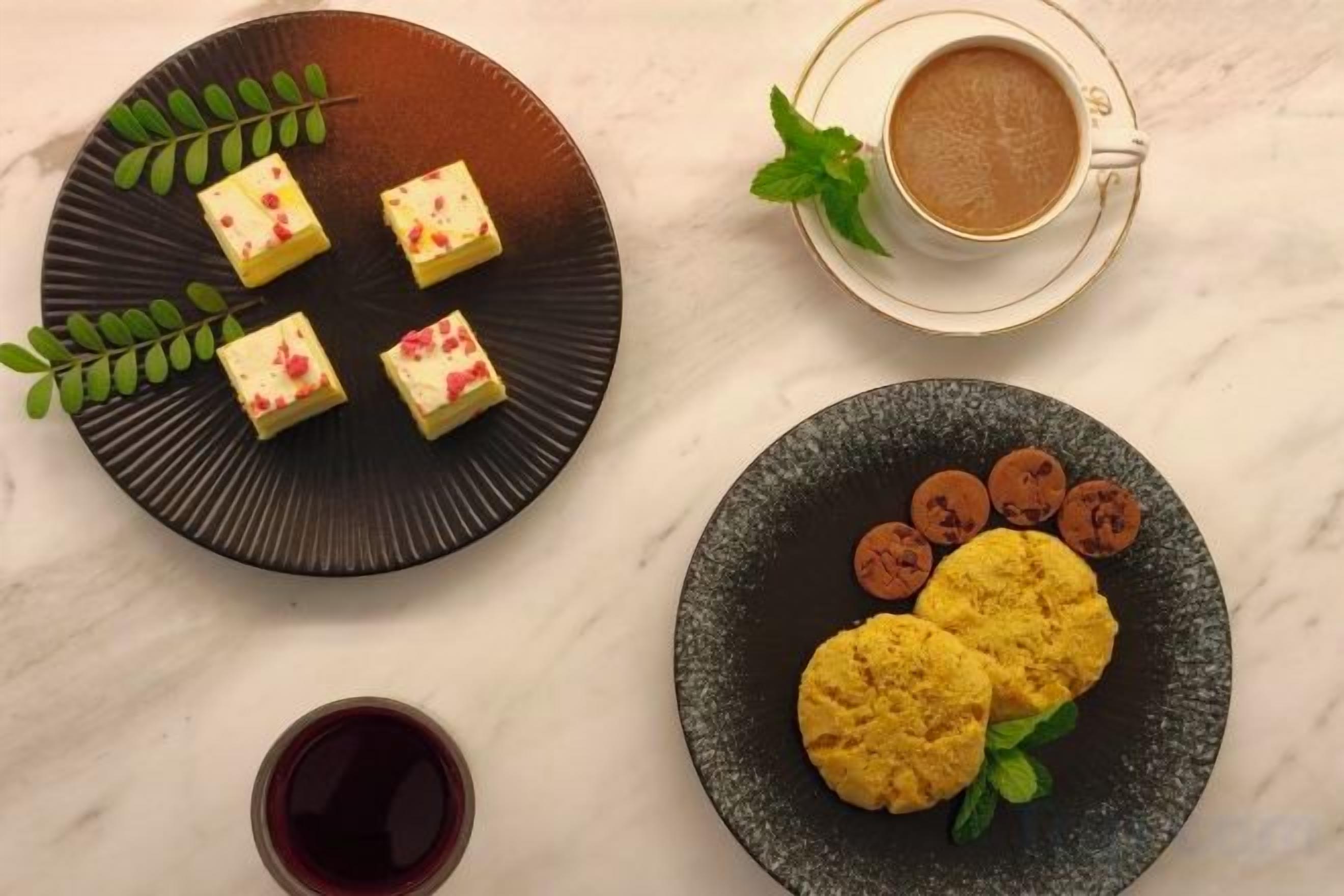 Una mesa con un plato de postres cuadrados, un plato de galletas, una taza de café y un vaso de bebida roja.