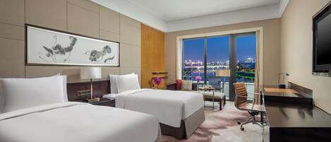 A hotel room with two beds, a desk, a chair, a TV, and a large window with a city view.