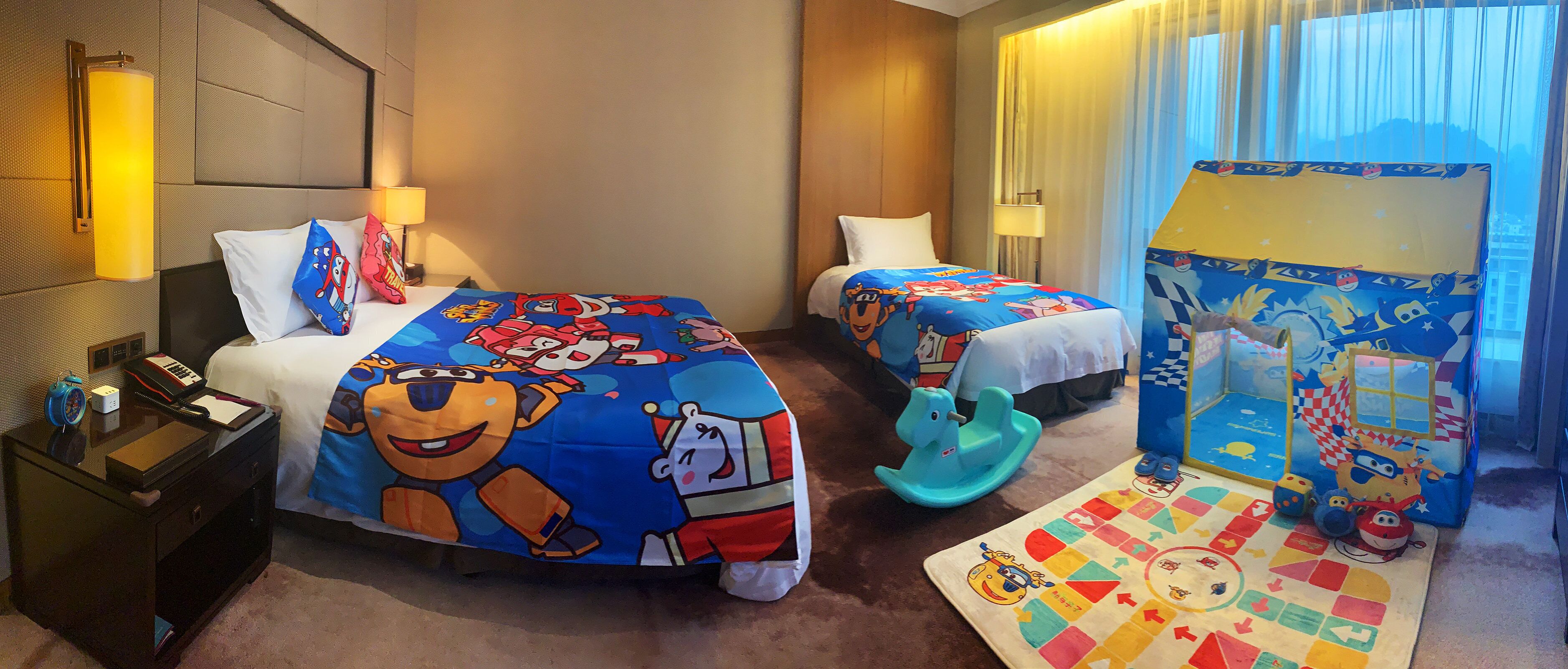 A hotel room with two beds, each with a colorful bedspread featuring cartoon characters. There is a desk with a lamp, a telephone, and a small clock. A play area with a mat and toys is visible in the corner.