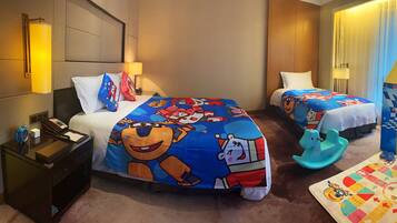 A hotel room with two beds, each with a colorful bedspread featuring cartoon characters. There is a desk with a lamp, a telephone, and a small clock. A play area with a mat and toys is visible in the corner.