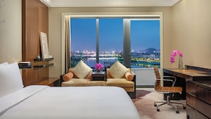 A hotel room with a large bed, two armchairs, a desk, and a view of a cityscape at night.