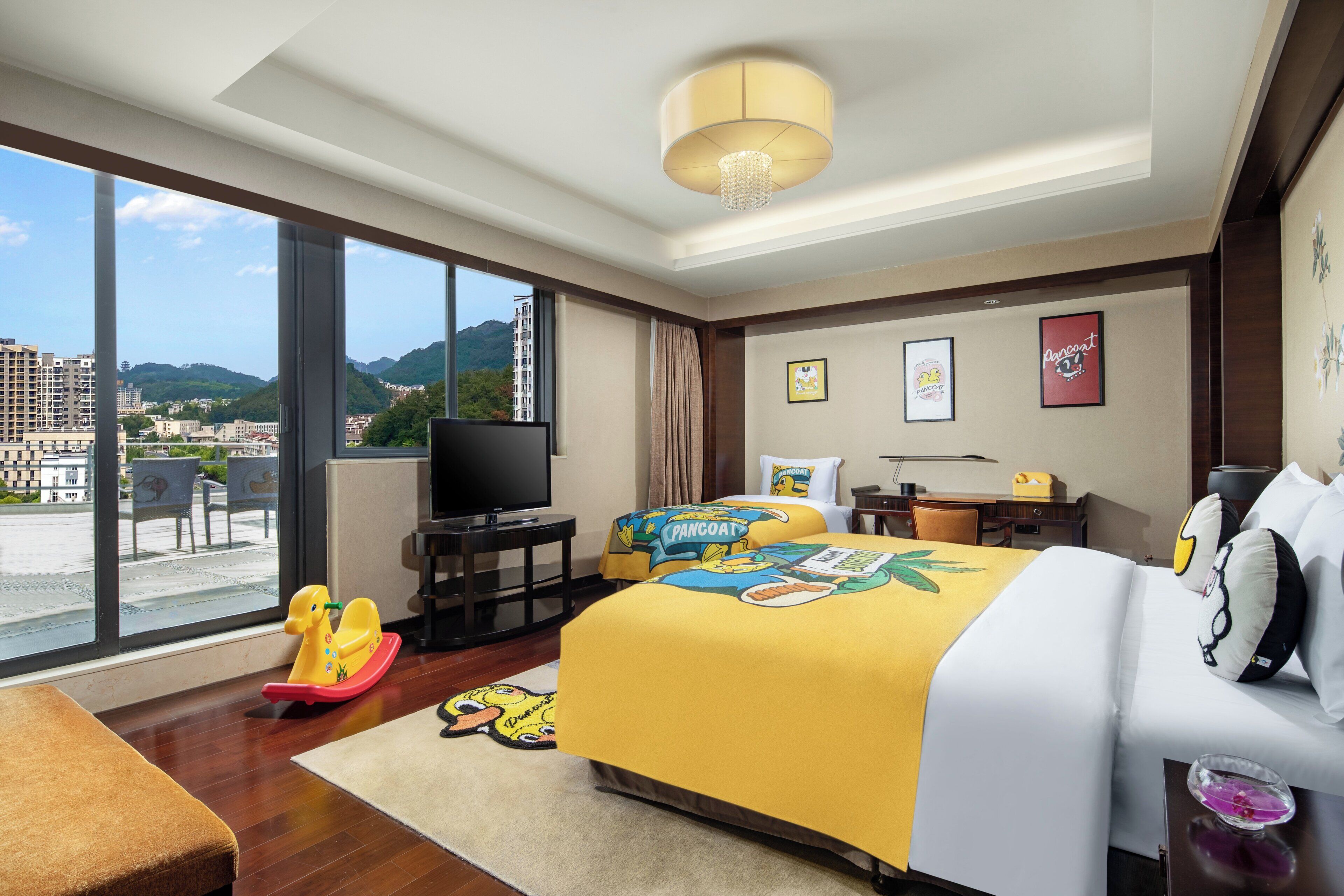A hotel room with a large window, a flat-screen TV, a bed with a yellow bedspread, a desk with a lamp, and a chair.