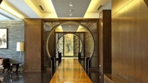 A modern hotel lobby with a long, polished wooden reception desk, a seating area with chairs, and a decorative wall with a circular design.