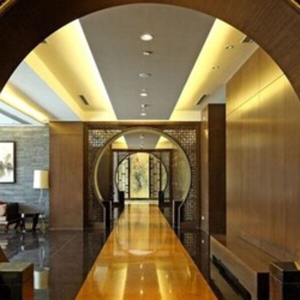 A modern hotel lobby with a long, polished wooden reception desk, a seating area with chairs, and a decorative wall with a circular design.