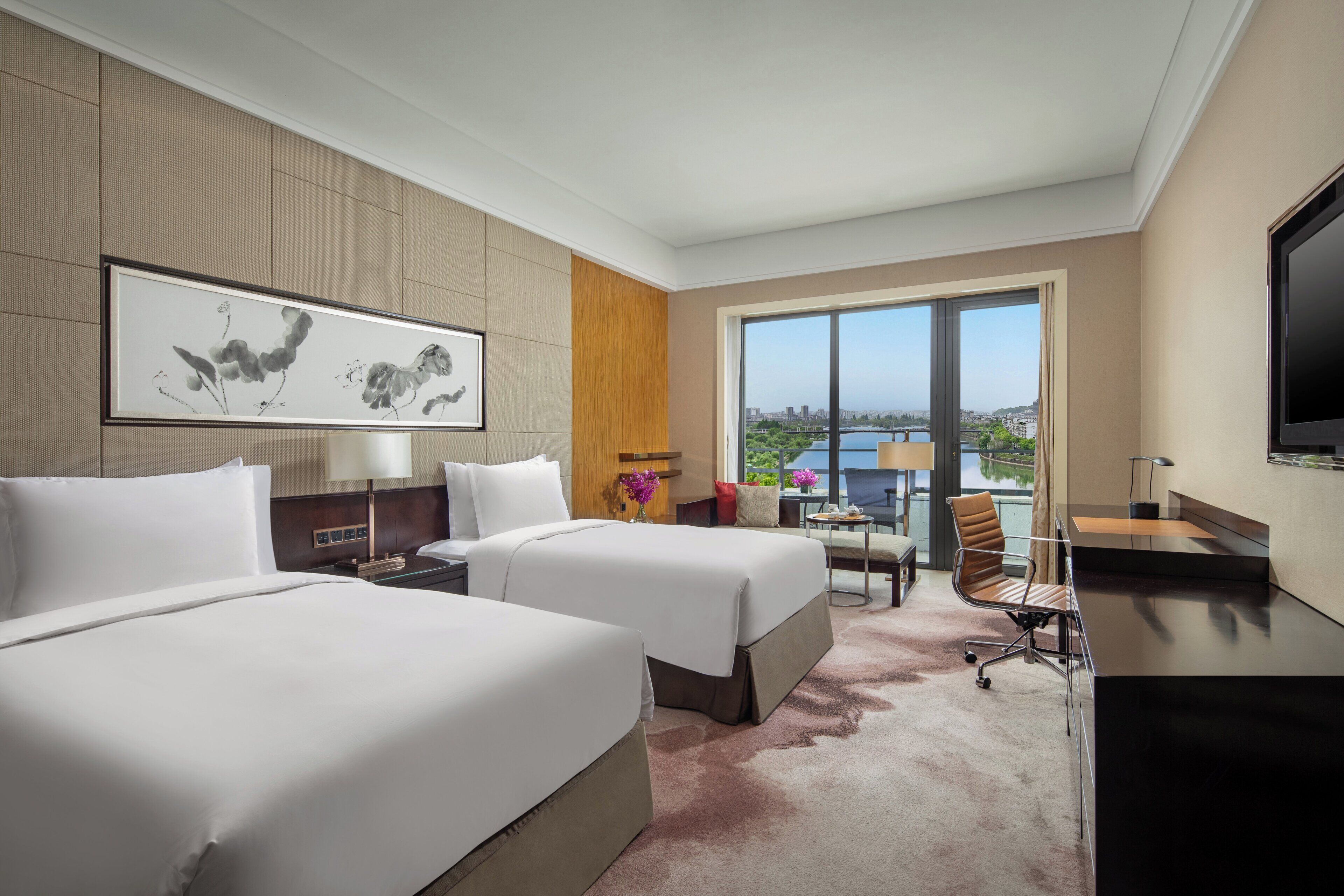 Ein Hotelzimmer mit zwei Betten, einem Schreibtisch, einem Stuhl, einem Fernseher und einem großen Fenster mit Blick auf die Stadt.