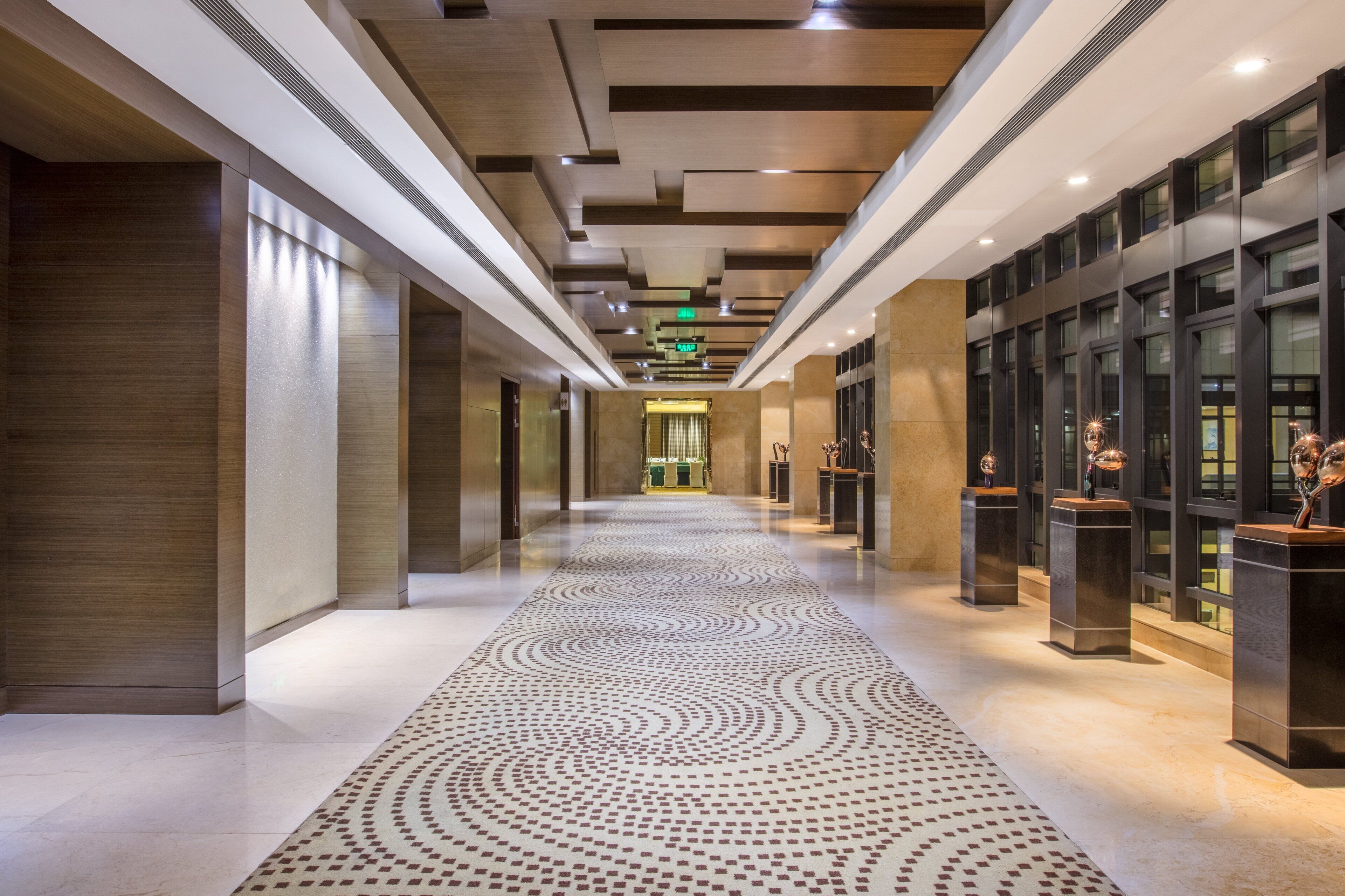 A modern hotel corridor with a patterned floor, recessed lighting, and a view into a room with a green table and chairs.