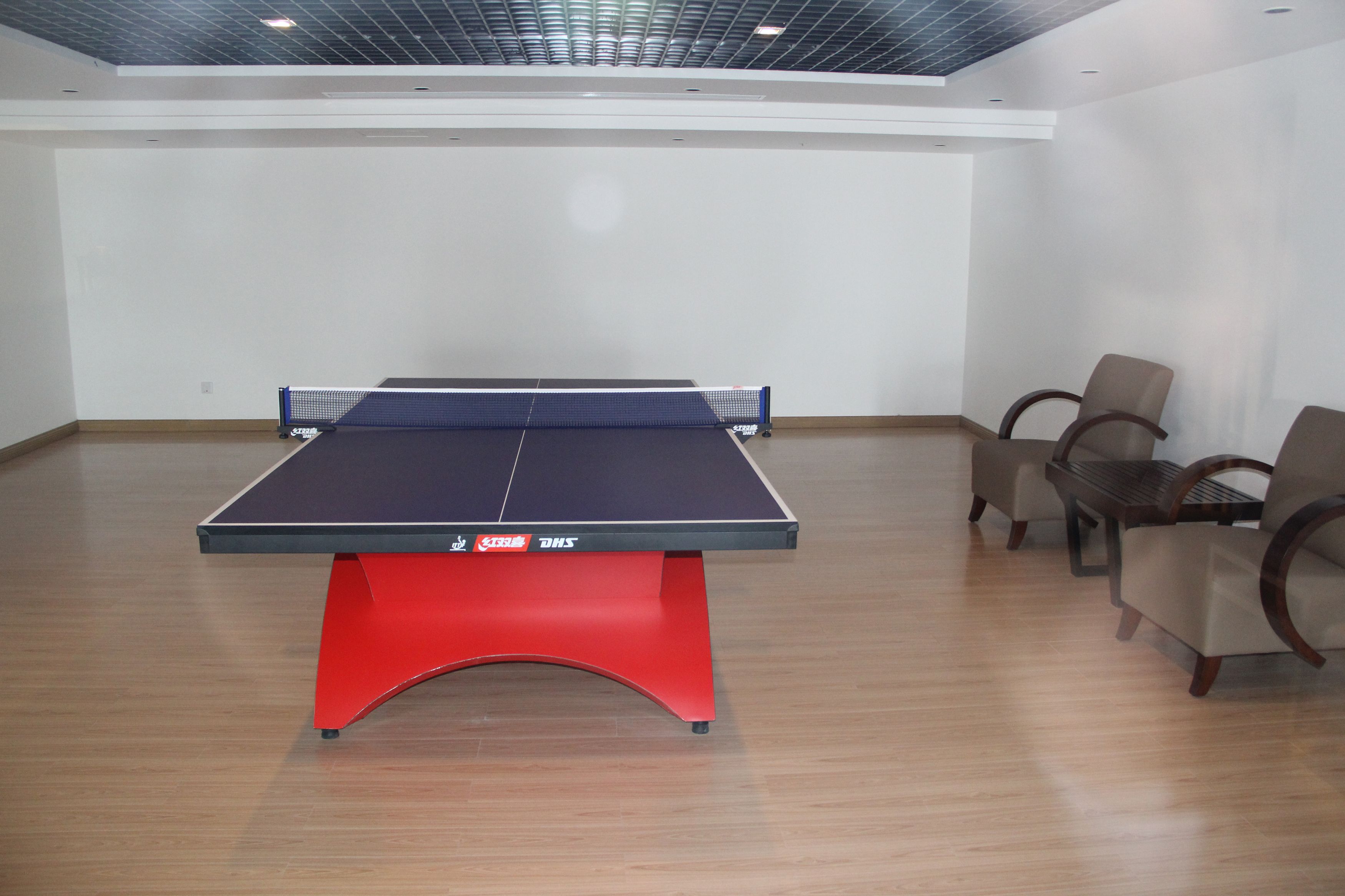A ping pong table with red legs and a blue playing surface, surrounded by four chairs with wooden frames and cushioned seats.