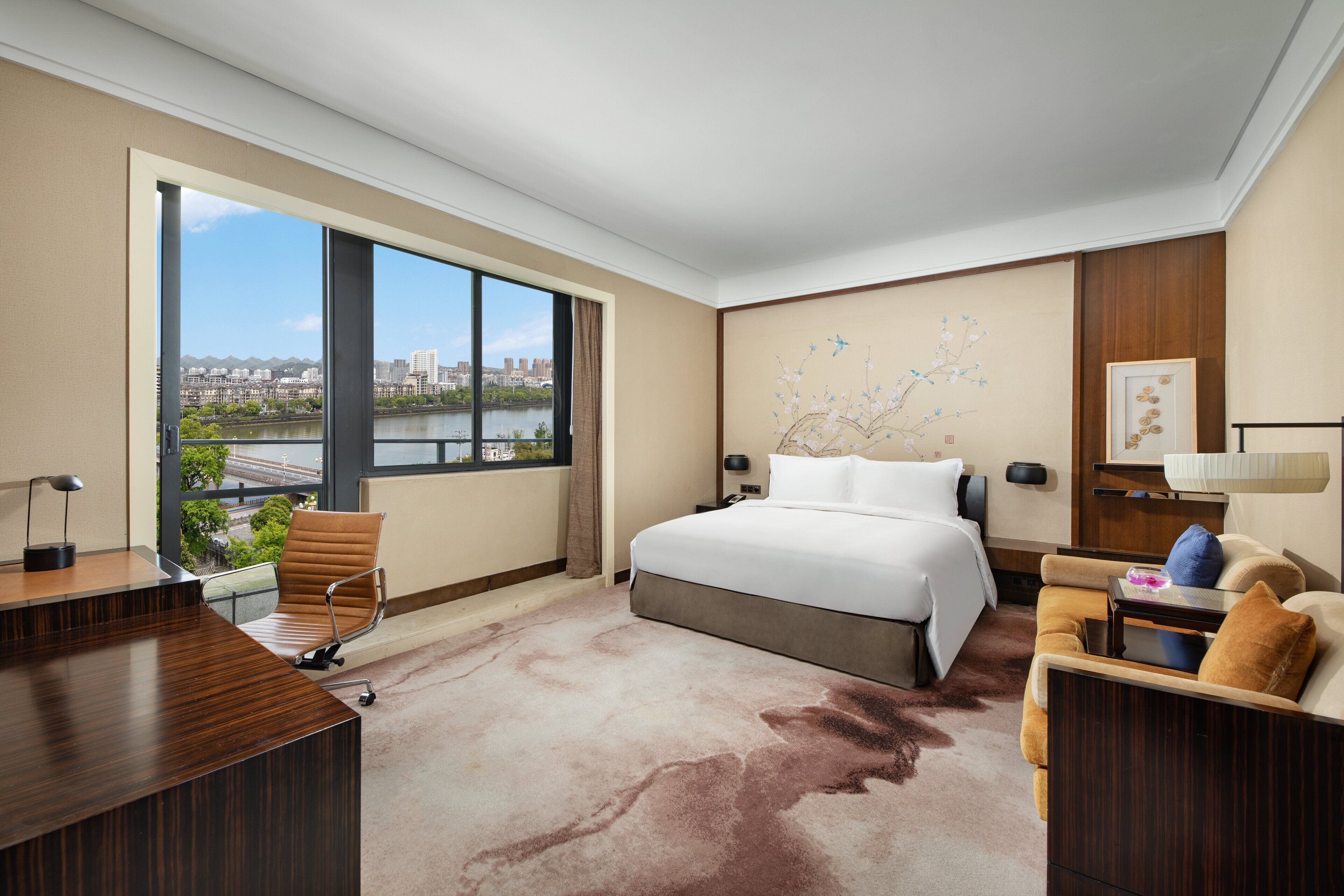A hotel room with a large bed, a desk, a chair, and a view of a cityscape through a large window.