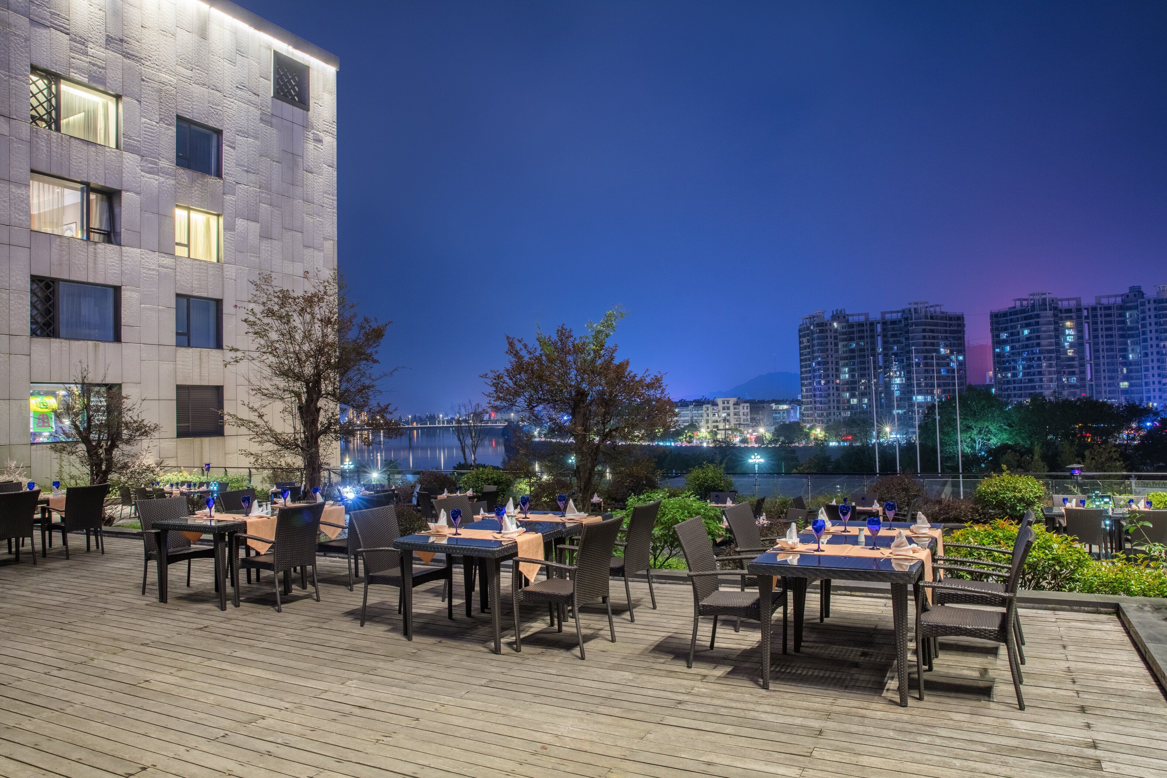 An outdoor dining area with tables and chairs, a view of a river, and buildings in the background.