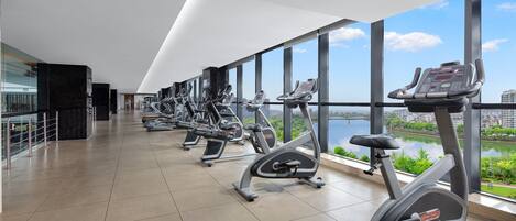 A modern gym with multiple stationary bikes and treadmills, large windows offering a view of a river and cityscape.