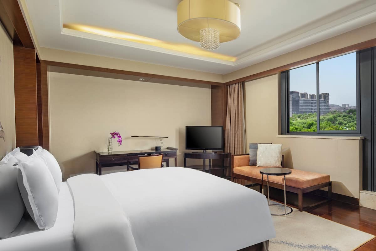 A modern hotel room with a large bed, a desk with a computer, a television, and a view of the city through large windows.
