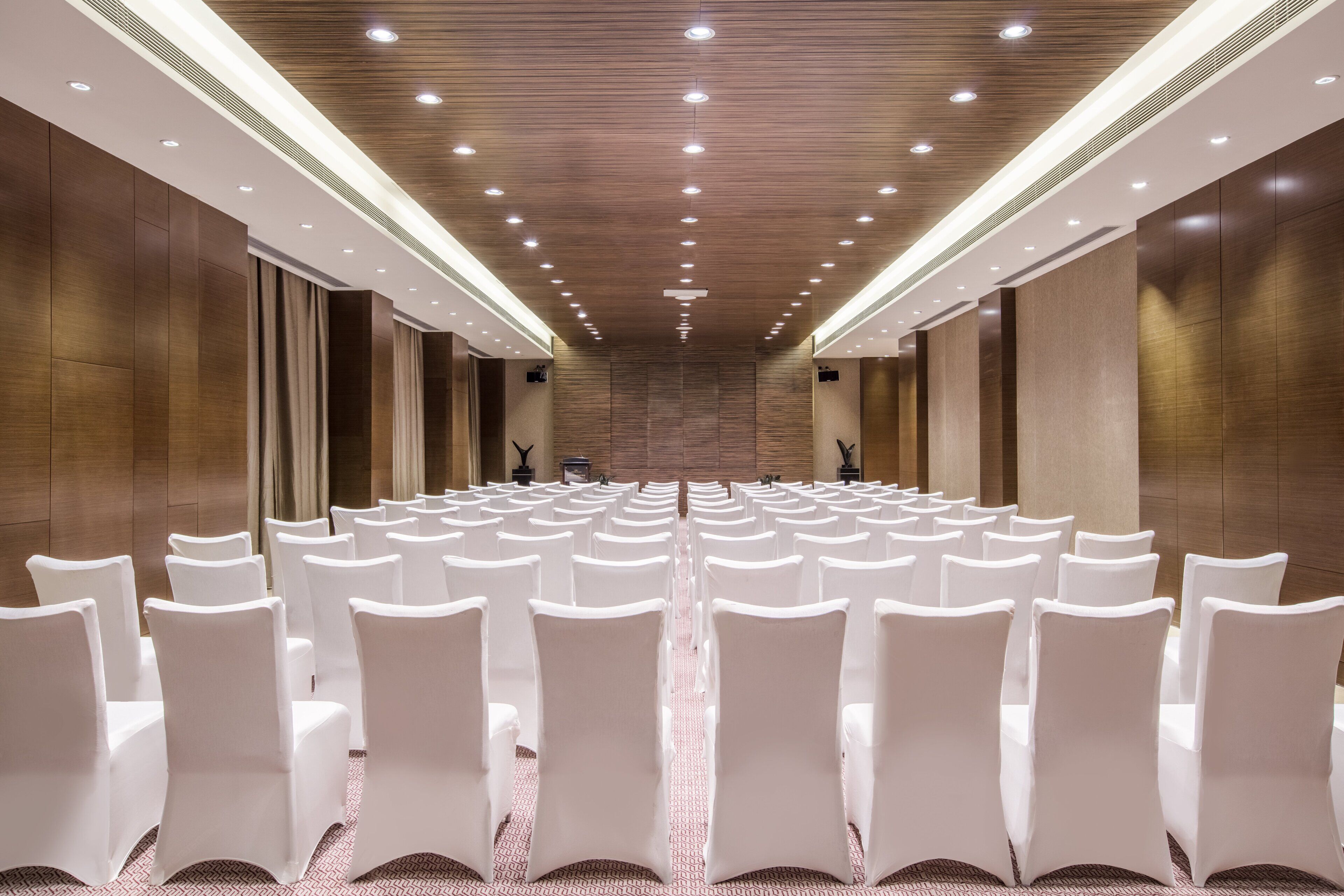 A conference room with rows of white chairs, wooden panel walls, and recessed lighting.