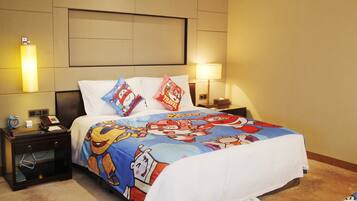 A hotel room with a bed featuring cartoon character bedding, a bedside table with a lamp, and a nightstand with a phone and clock.