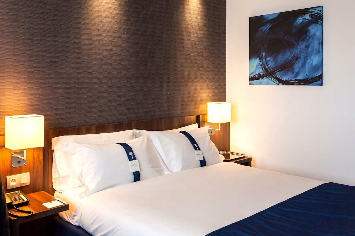 A hotel room with a wooden headboard, a large bed with white linens, two bedside lamps, and a painting on the wall.