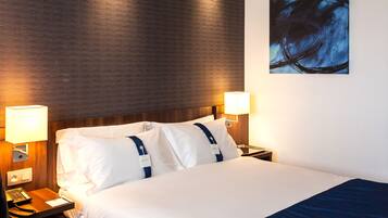 Una habitación de hotel con cabecera de madera, una cama grande con ropa blanca, dos lámparas de noche y un cuadro en la pared.
