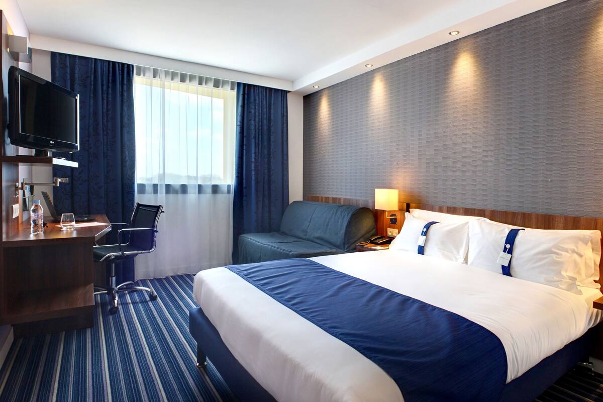 A hotel room with a large bed, a desk, a chair, a television, and a window with curtains.