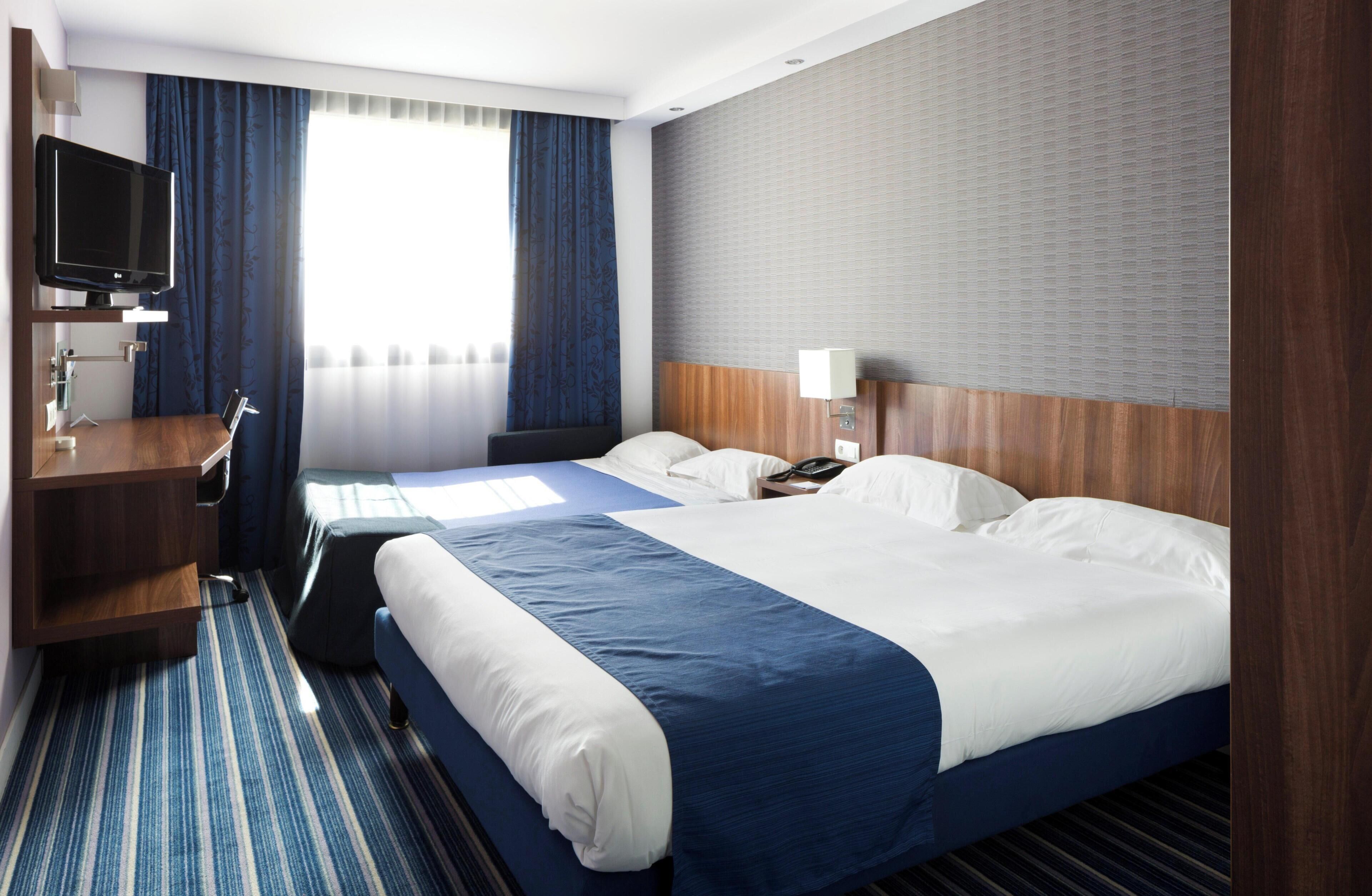 Une chambre d’hôtel avec un grand lit, un bureau, une télévision et une fenêtre avec des rideaux.
