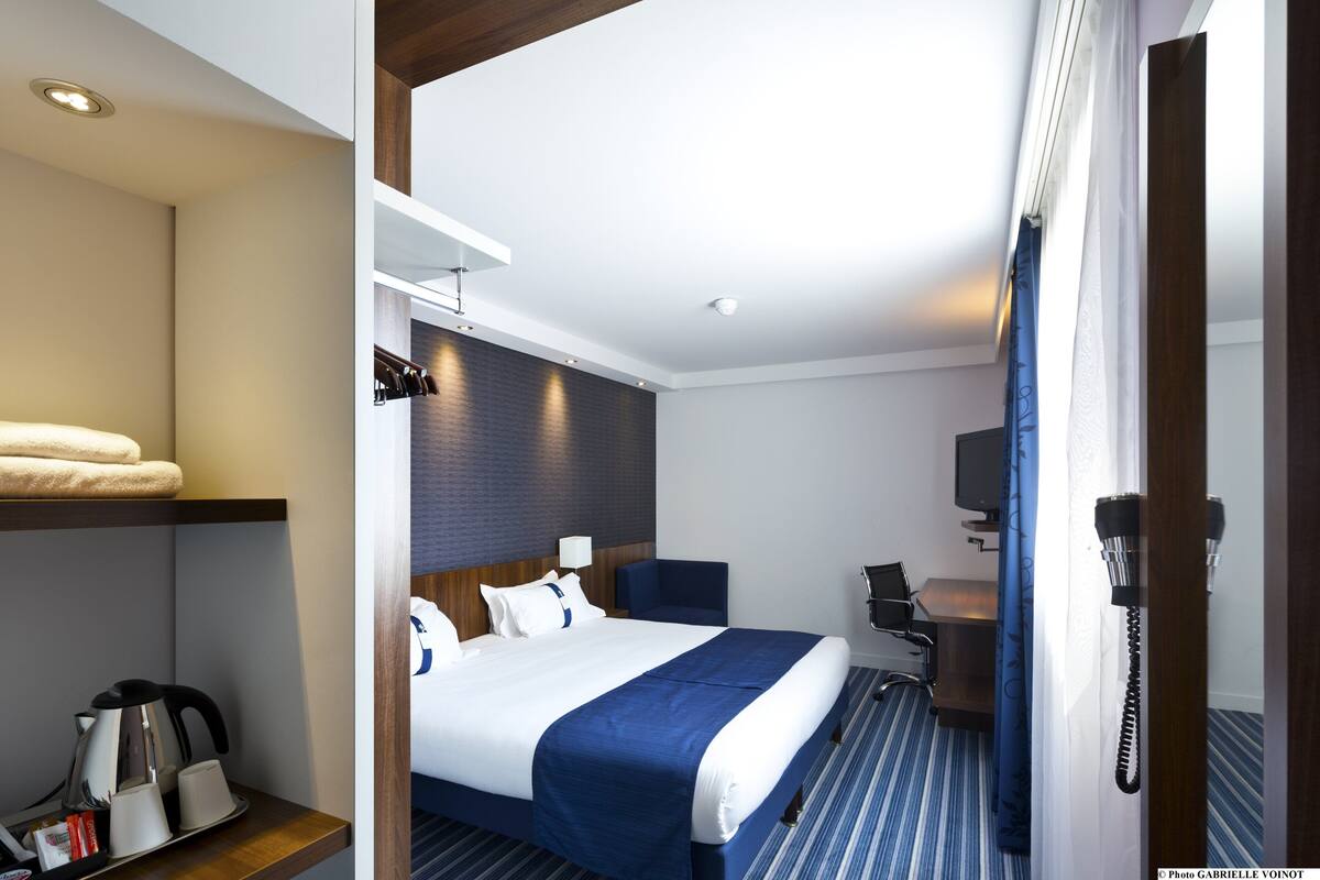 A hotel room with a bed, a desk, a chair, a telephone, and a shelf with towels and a kettle.