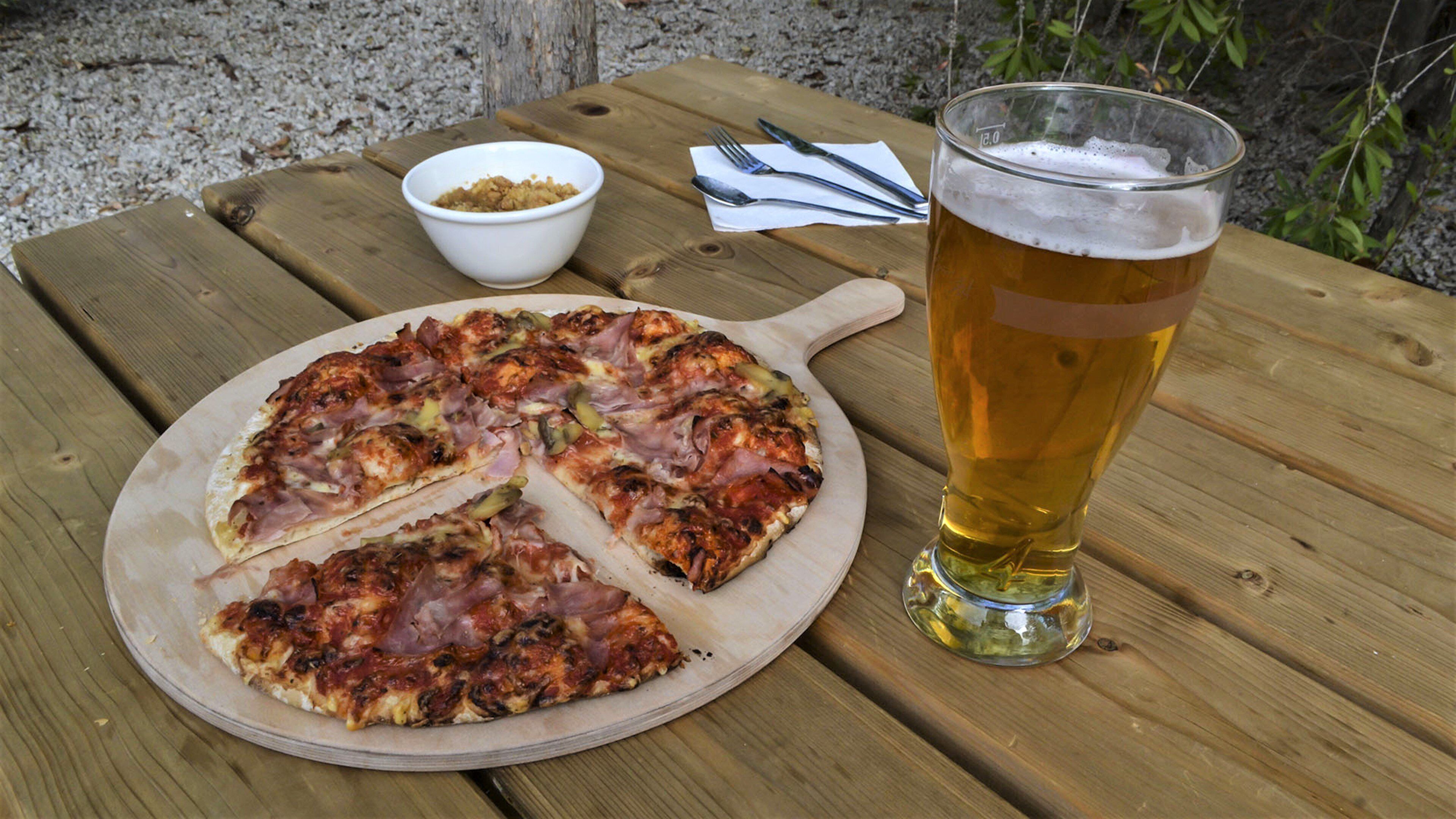 A pizza on a wooden board, a glass of beer, and a small bowl of sauce on a wooden table.