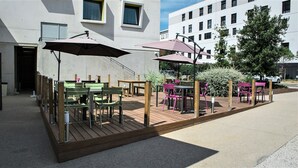 An outdoor patio with wooden tables and chairs, umbrellas, and a view of a modern building in the background.