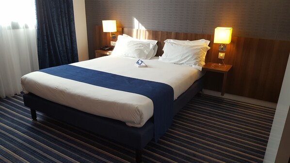 Una habitación de hotel con una cama grande, dos lámparas de noche, un cabecero de madera y una alfombra a rayas.