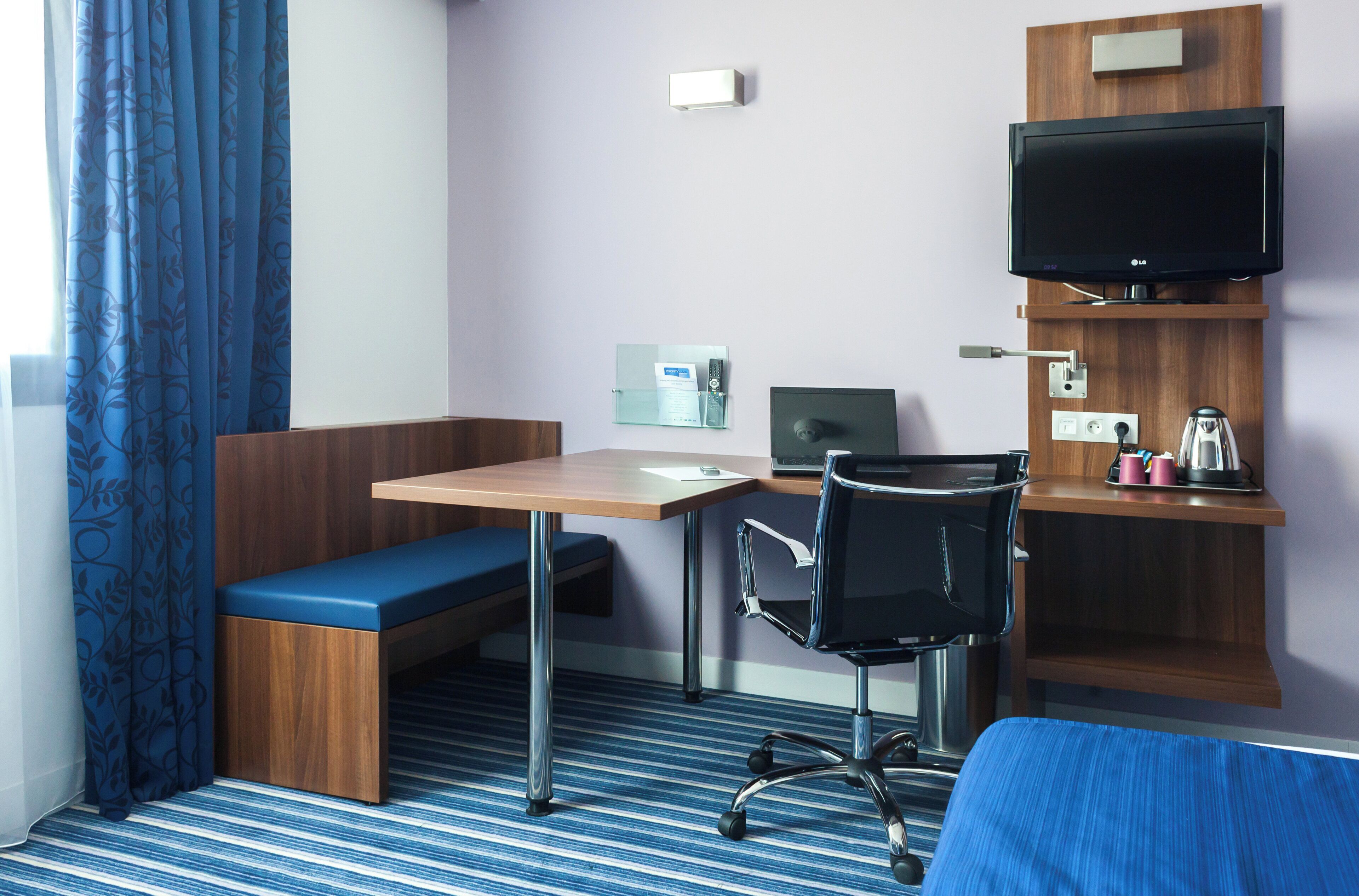 A hotel room with a desk, chair, bench, television, and a small shelf with items.