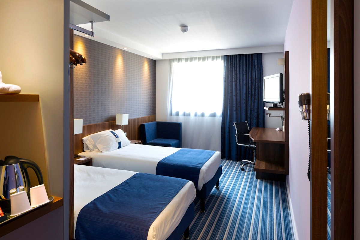 A hotel room with two beds, a desk, a chair, a TV, and a coffee machine.