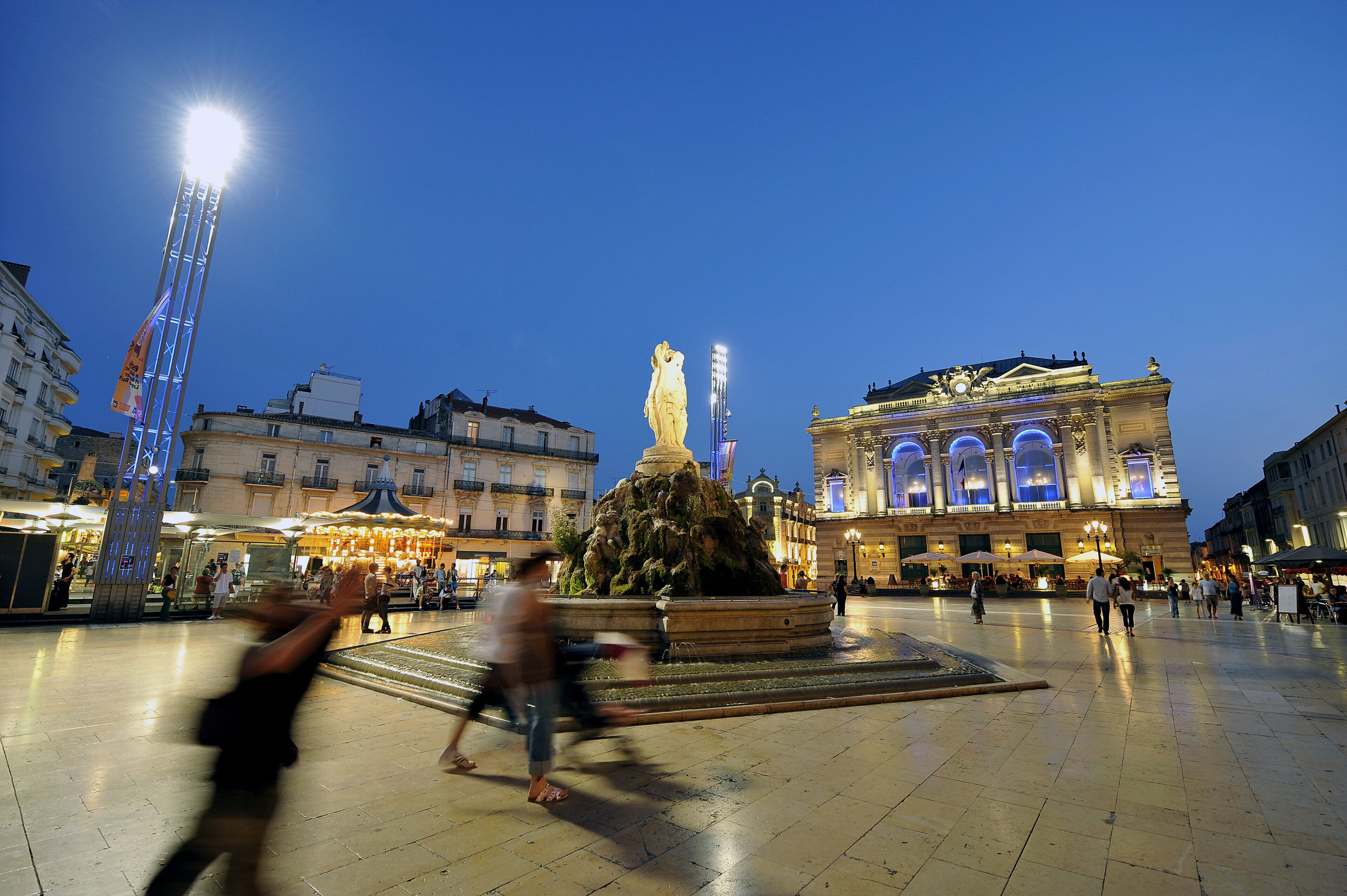 Una plaza pública con una fuente, edificios y un campanario iluminado.