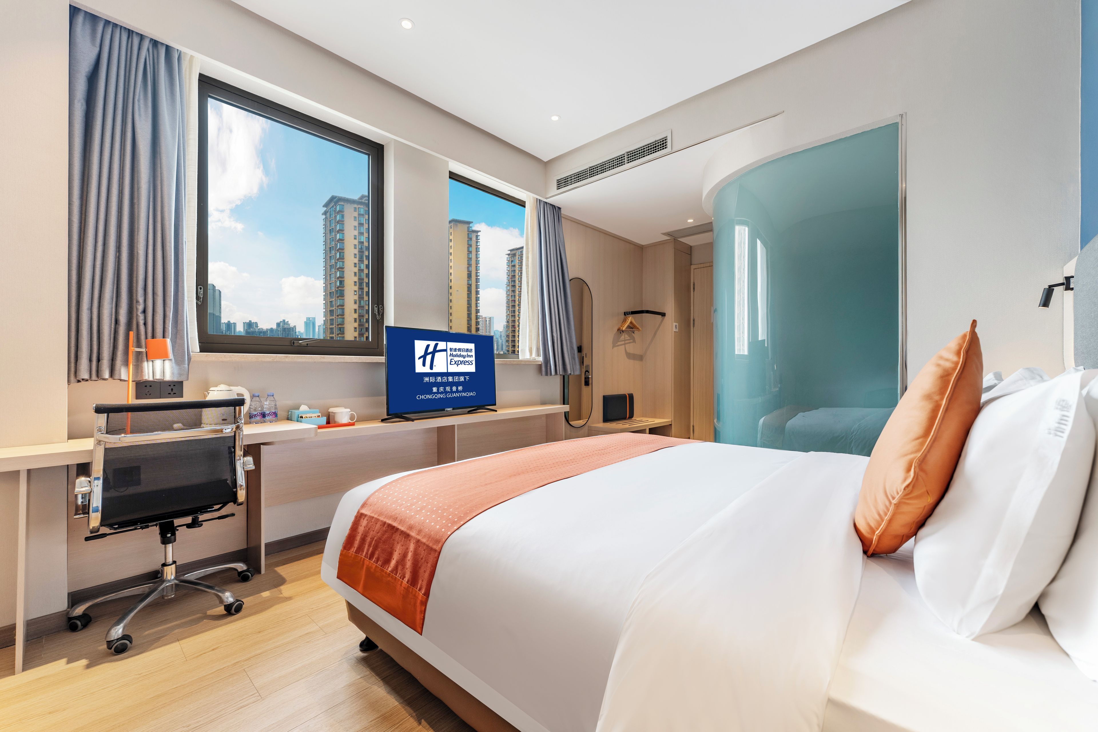 A hotel room with a large bed, a desk with a computer, a window with curtains, and a view of tall buildings.