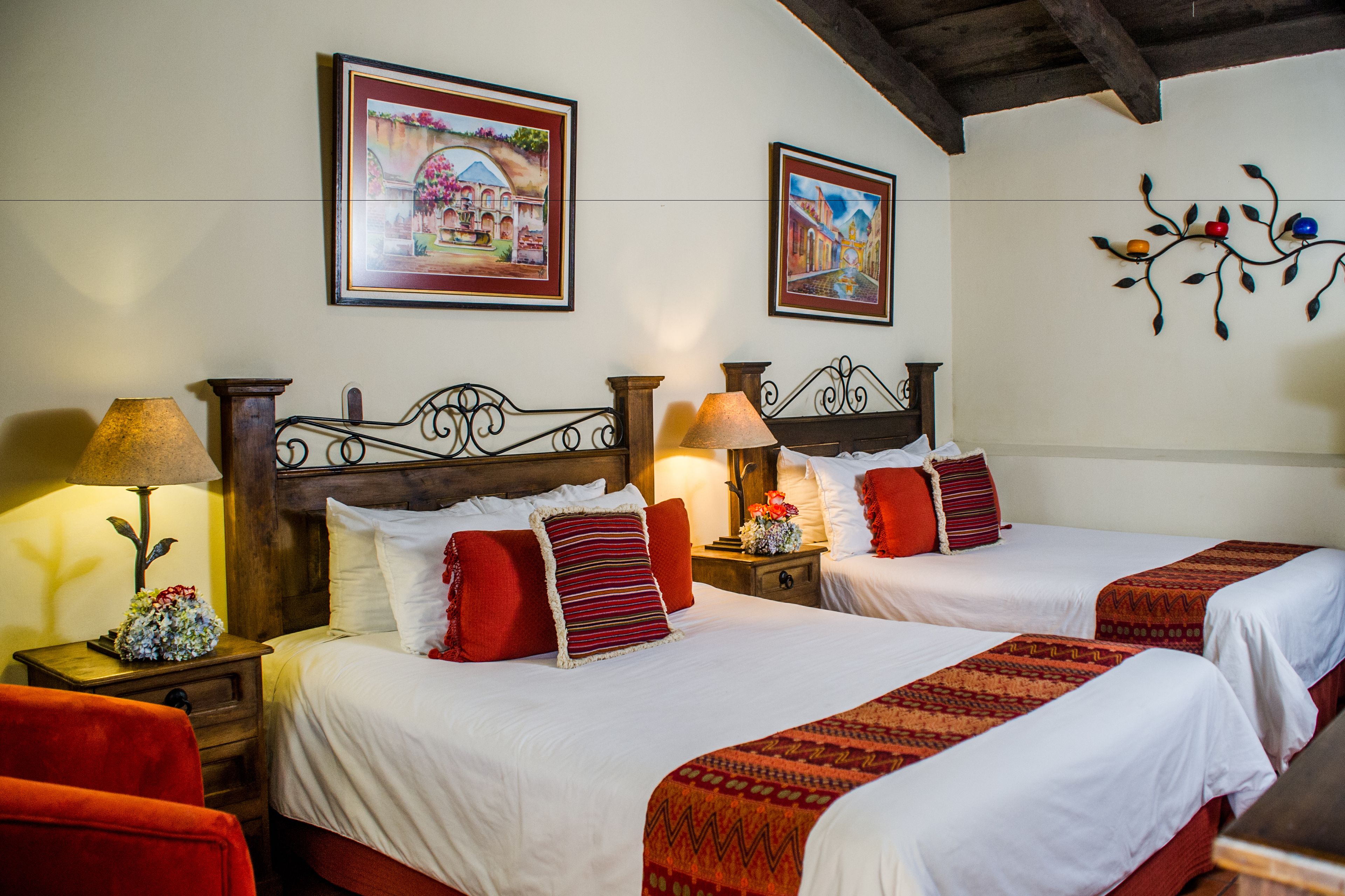 A hotel room with two beds, each with a wooden headboard, white bedding, and red accents. There are two bedside tables with lamps and floral arrangements.