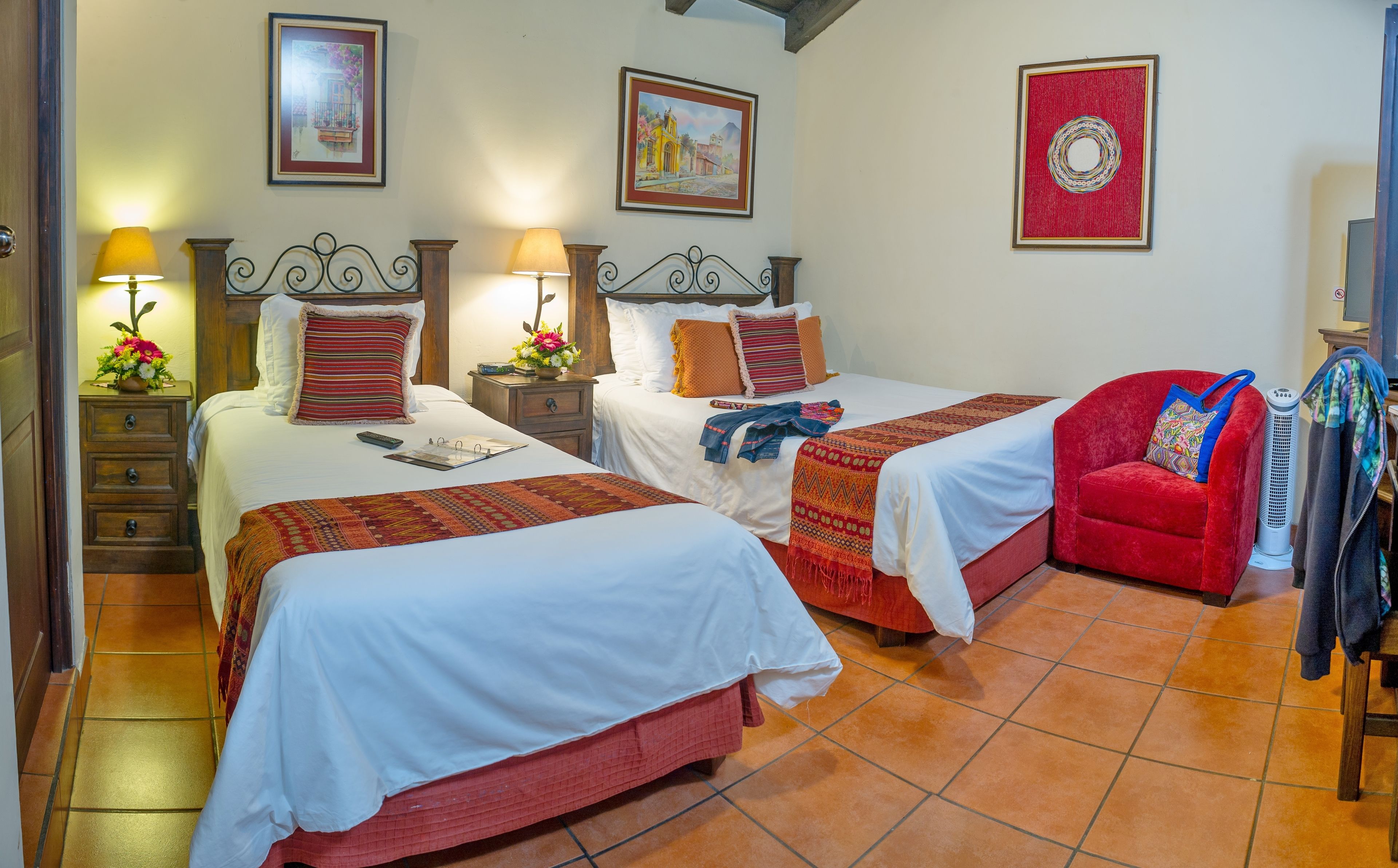 A hotel room with two beds, a red armchair, a heater, and artwork on the walls.