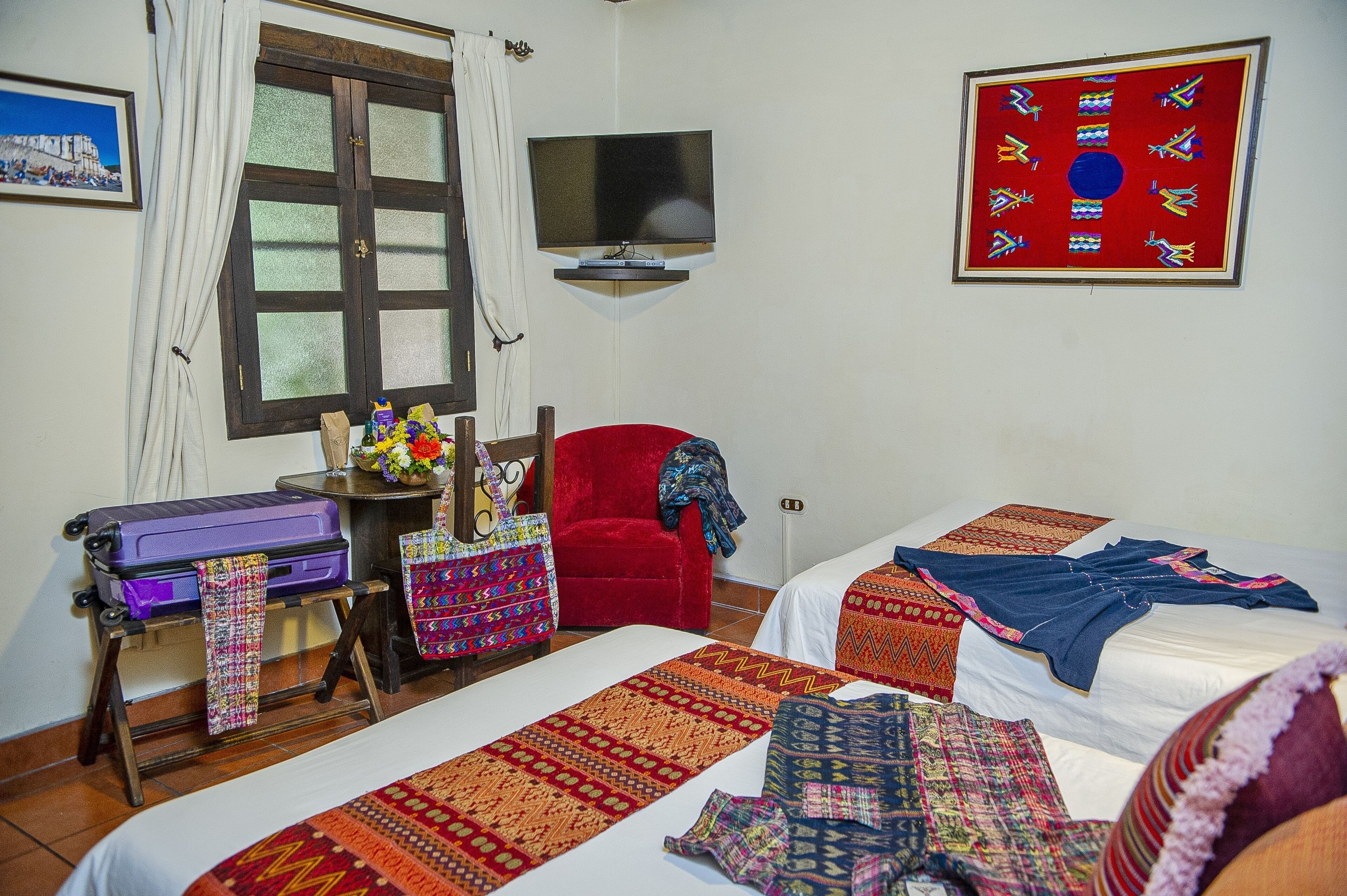 A hotel room with two beds, a TV mounted on the wall, a window with curtains, a suitcase, and a table with a vase of flowers.