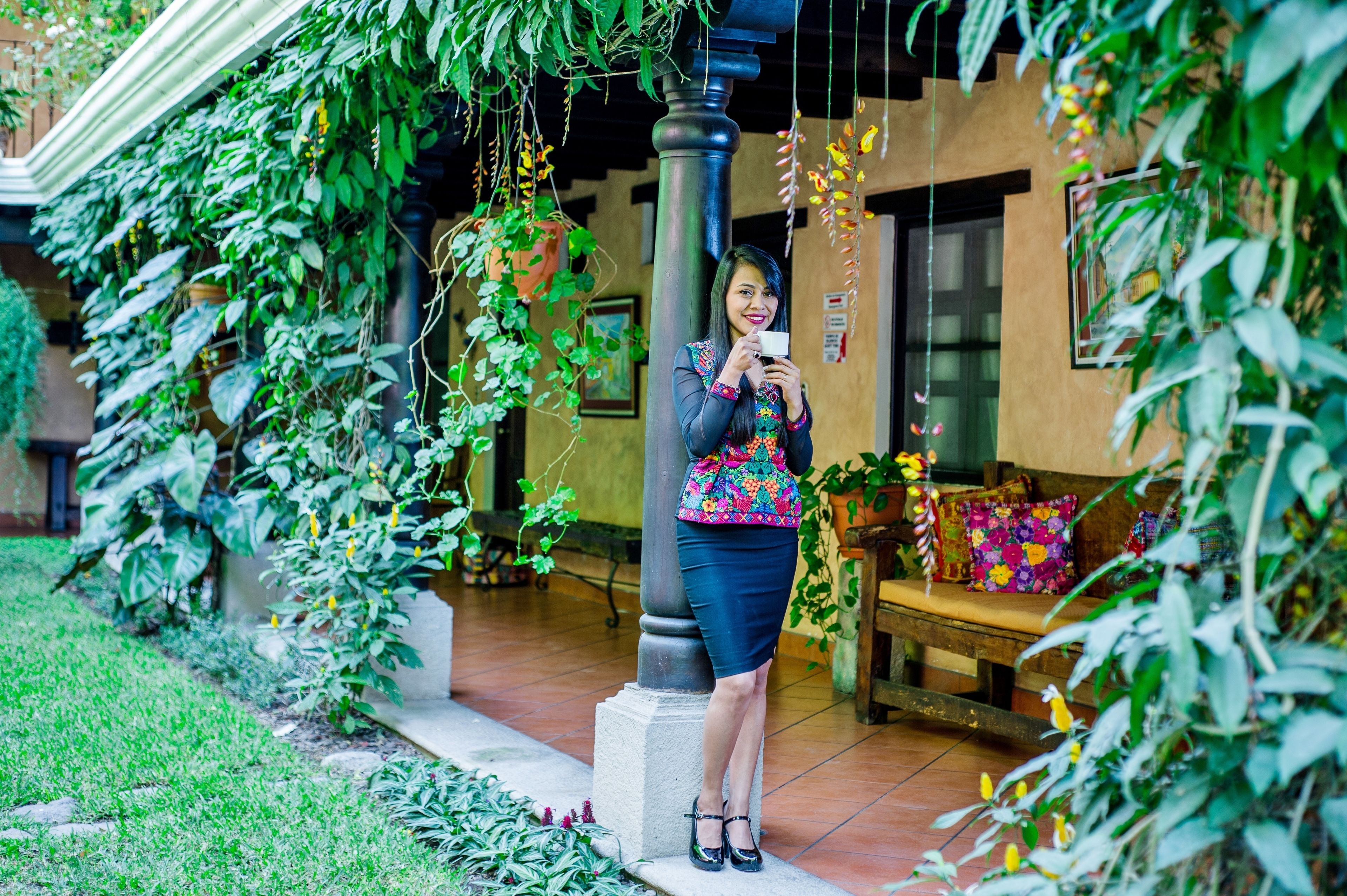 A person standing in a garden area with hanging plants and a bench with colorful cushions.