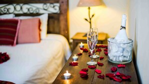 A hotel room with a neatly made bed, a bedside table with a bottle of champagne, a glass, and ice cubes, and a lit candle.
